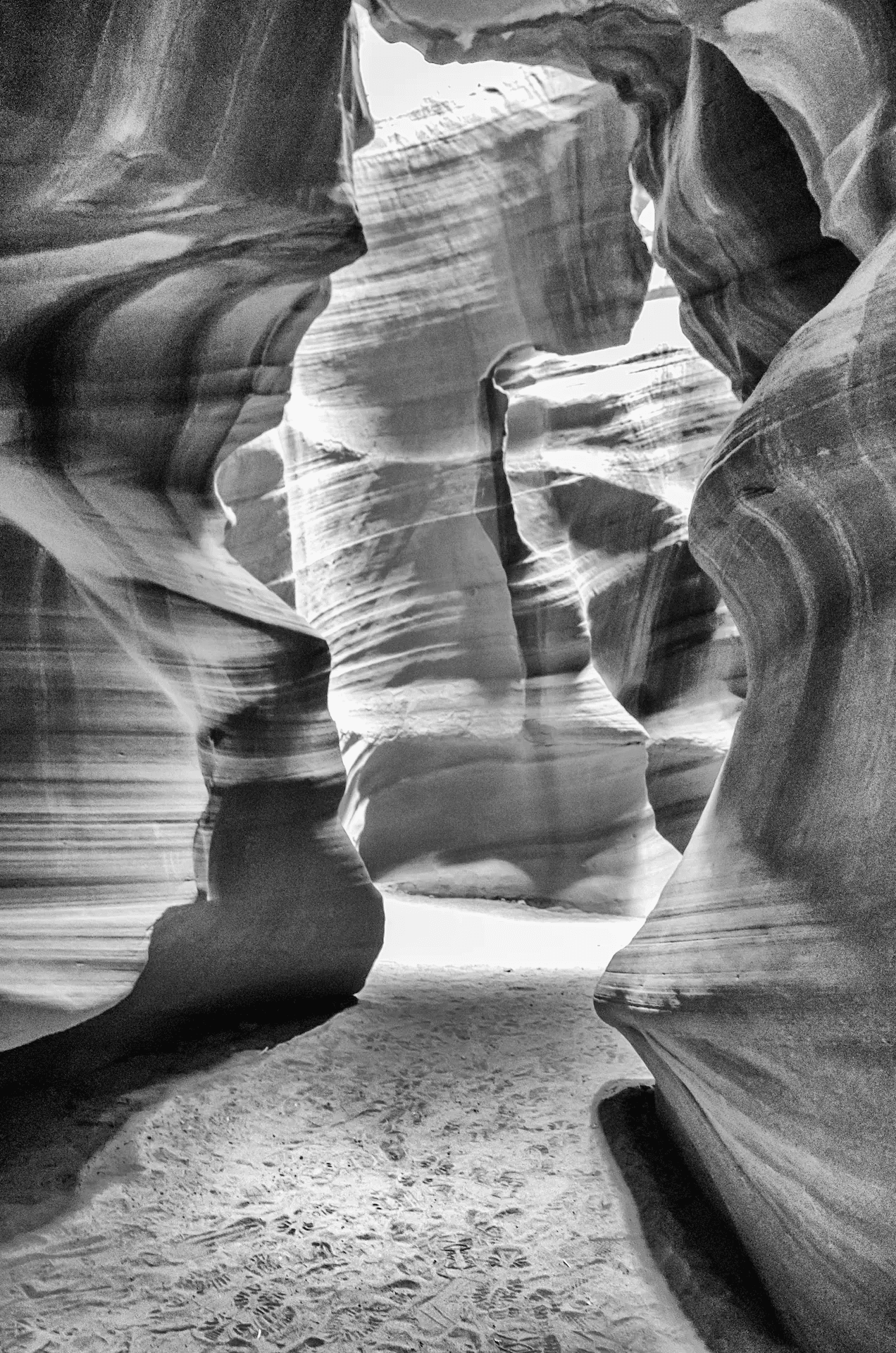 Black and white landscape photograph of Antelope Canyon, Arizona. The image captures the interior of a slot canyon, showcasing the wavy, sculpted sandstone walls in shades of grey. Light and shadow emphasize the textures and contours of the canyon walls. Atmospheric monochrome nature photography. 