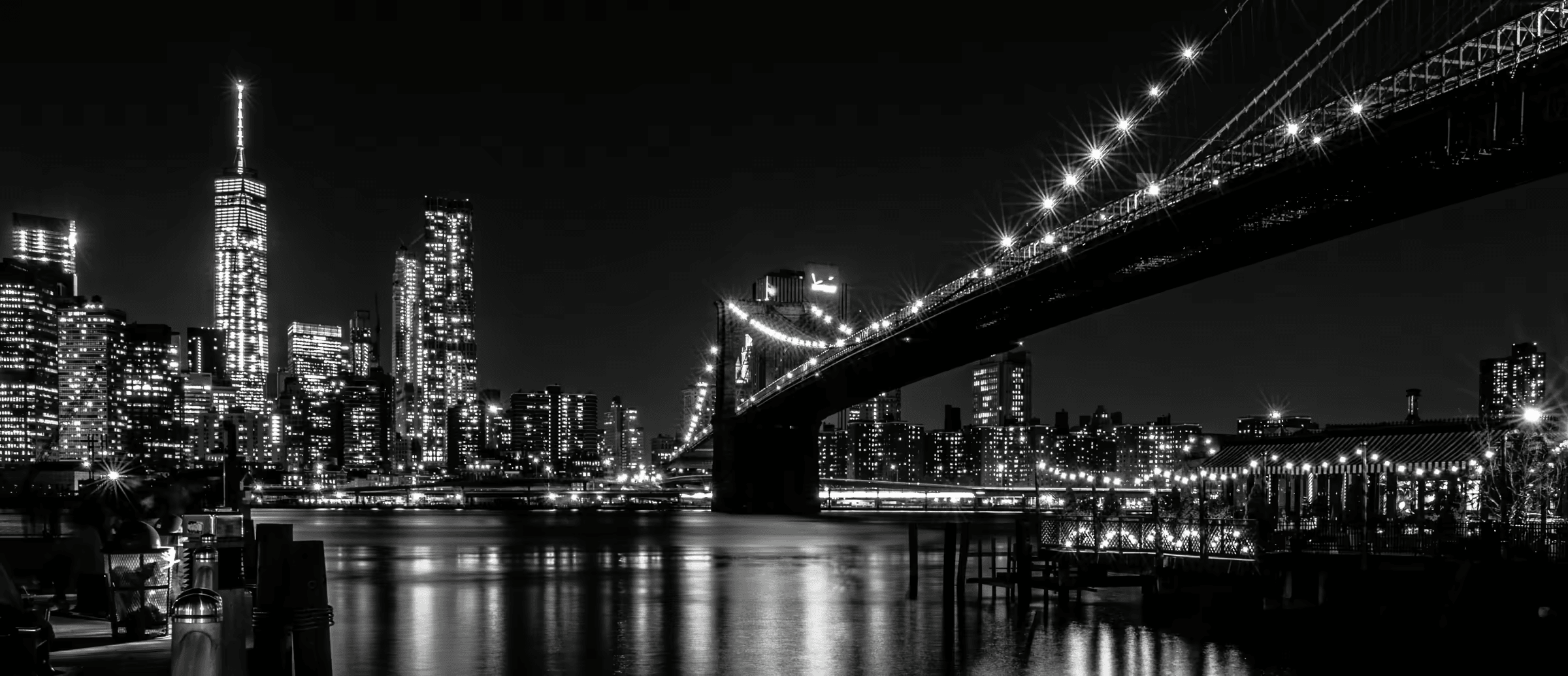 Black and white cityscape photograph of the Brooklyn Bridge and Manhattan skyline at night. The Brooklyn Bridge dominates the right side of the frame, illuminated by string lights. The Manhattan skyline, punctuated by One World Trade Center, twinkles with city lights in the background. The East River reflects the city lights. Evocative monochrome urban landscape.