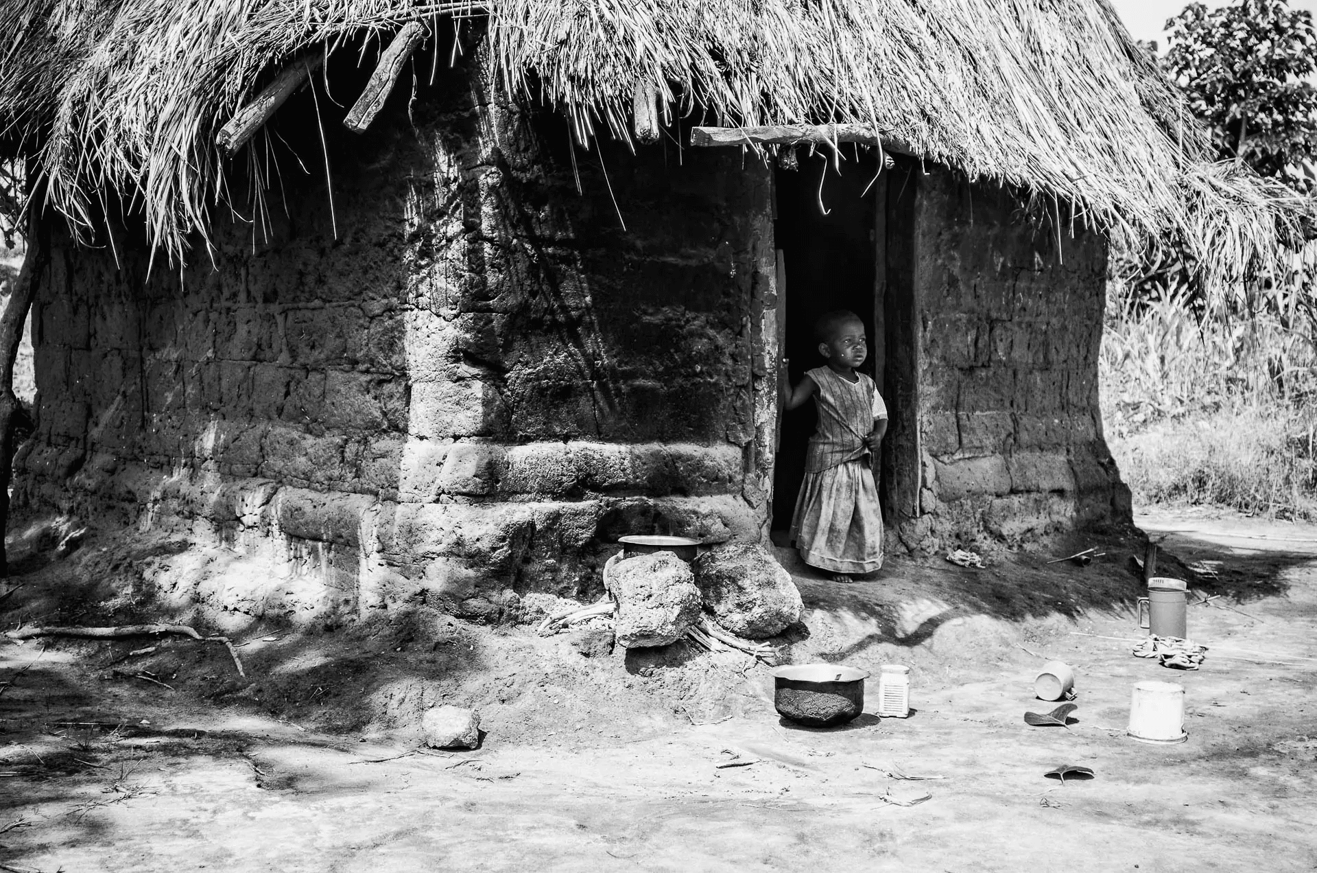 A black and white portrait photograph of a young African child standing in the doorway of a simple mud hut. The child, dressed in a patterned dress, looks thoughtfully out of frame. The hut is constructed of rough mud bricks and has a thatched roof.  Cooking utensils and containers are scattered on the dusty ground in front of the hut, suggesting a humble rural dwelling. The image is rich in texture and shadow, capturing a moment of quiet anticipation in a simple setting.