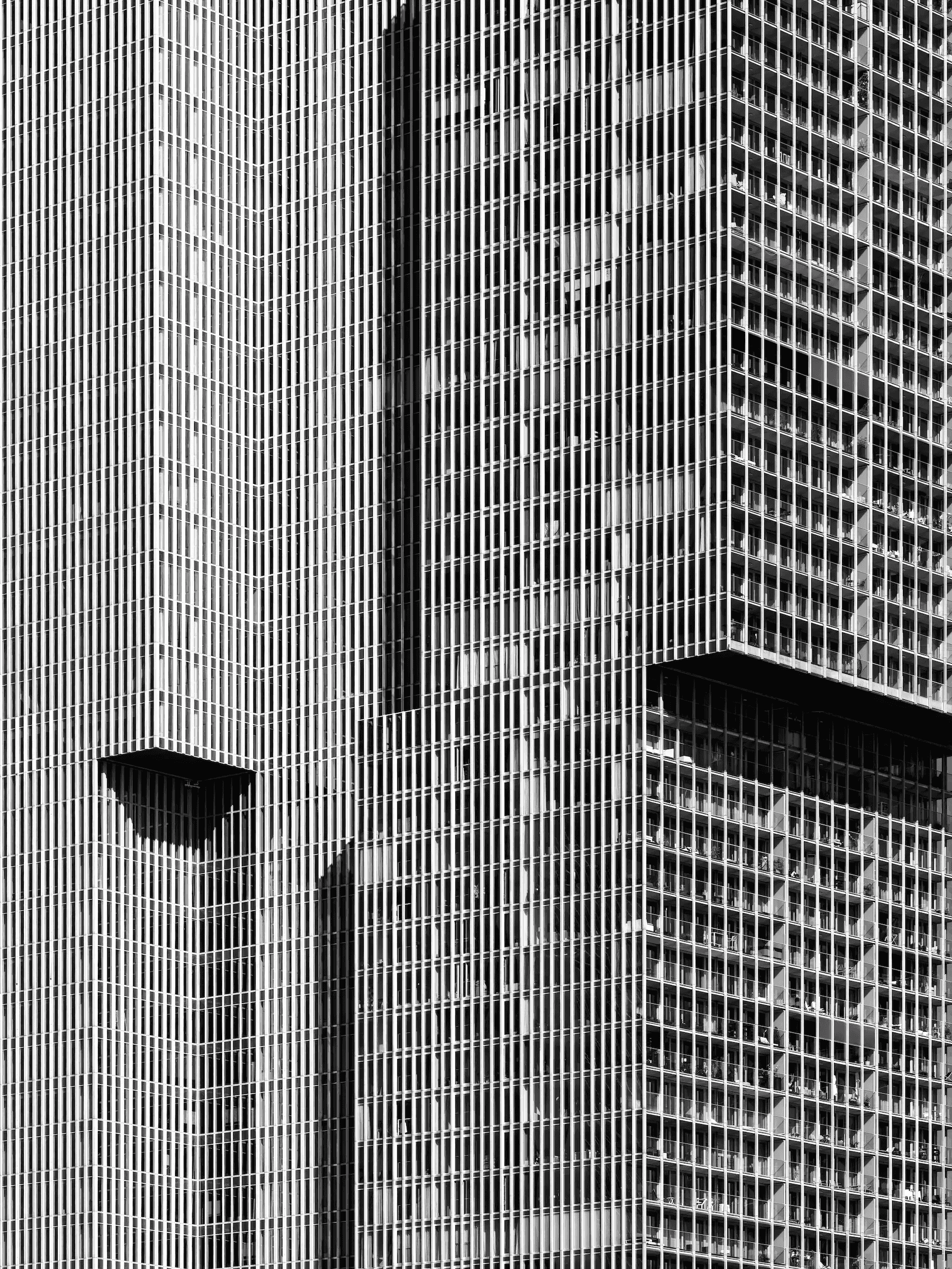 Abstract black and white photograph of the facade of the 'De Rotterdam' building. The image is a tight crop showing the repeating pattern of vertical lines and windows, creating a geometric and textured effect.