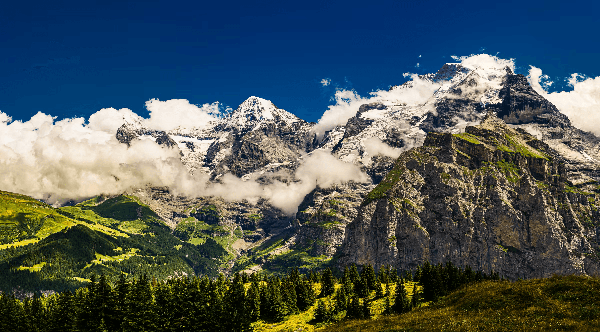 Landscape photograph of the Eiger, Mönch, and Jungfrau mountains in the Swiss Alps. The three peaks, partially shrouded in clouds, dominate the skyline against a deep blue sky. Lush green hills and forests cover the foreground slopes leading up to the imposing mountains. Daytime scenic view of the Swiss alpine landscape.