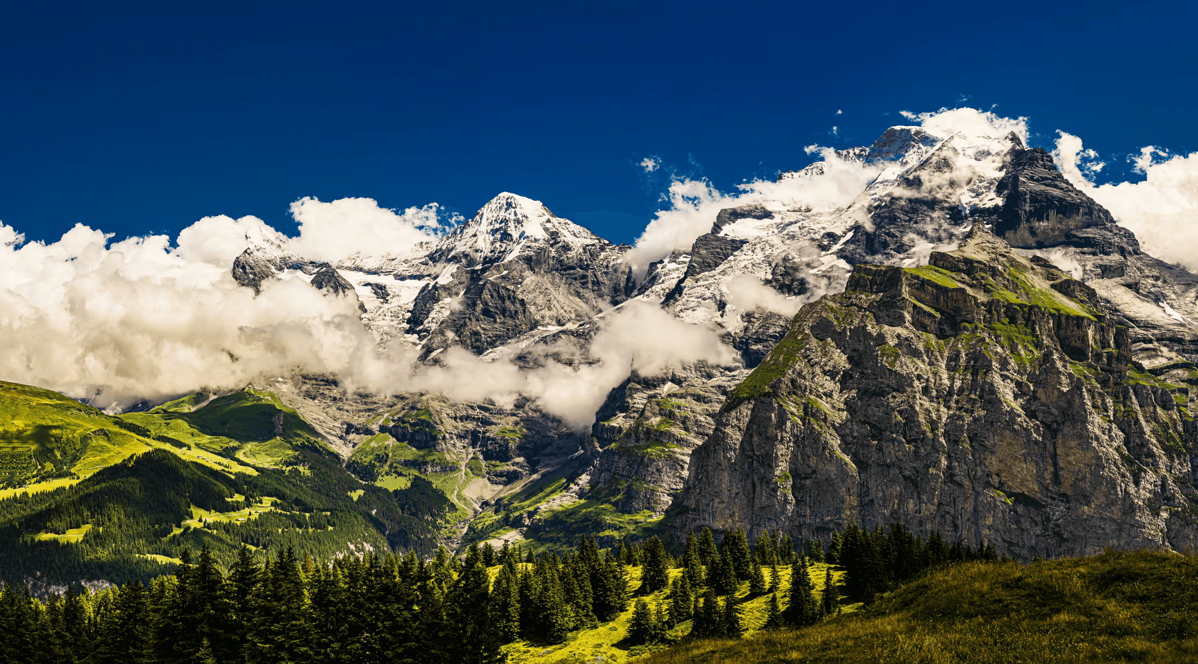 Landscape photograph of the Eiger, Mönch, and Jungfrau mountains in the Swiss Alps. The three peaks, partially shrouded in clouds, dominate the skyline against a deep blue sky.