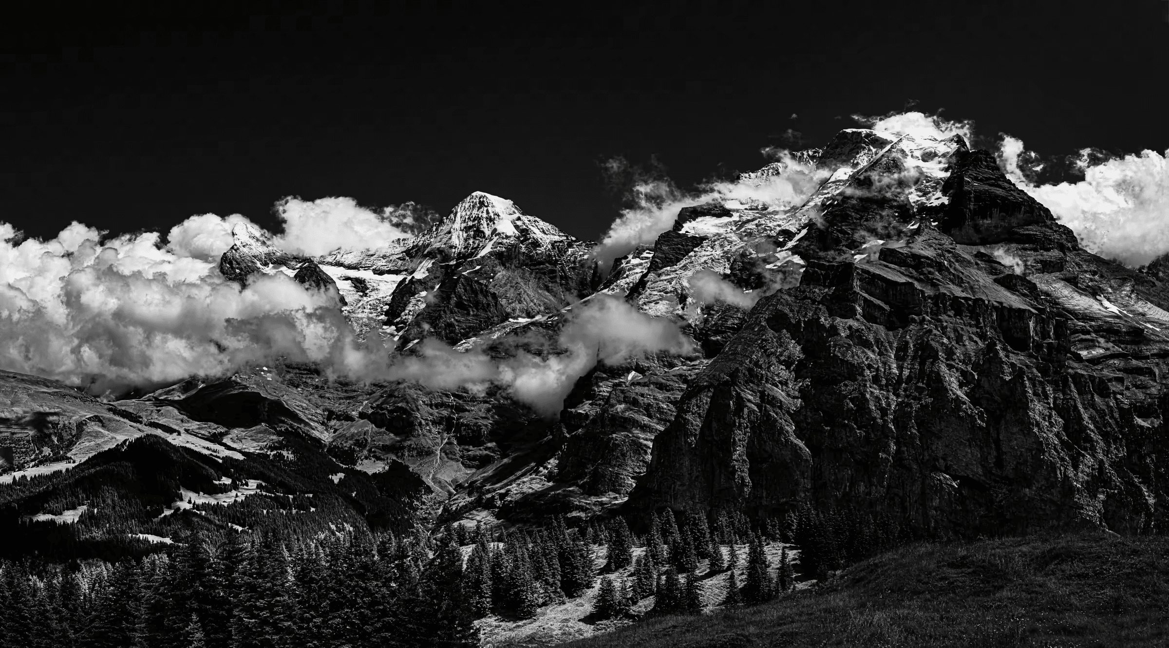 Black and white landscape photograph of the Eiger, Mönch, and Jungfrau mountains in the Swiss Alps, viewed from Mürren. The three peaks dominate the upper portion of the frame, partially obscured by dramatic clouds. The lower slopes are covered in dark forests. Evocative monochrome mountain scenery.