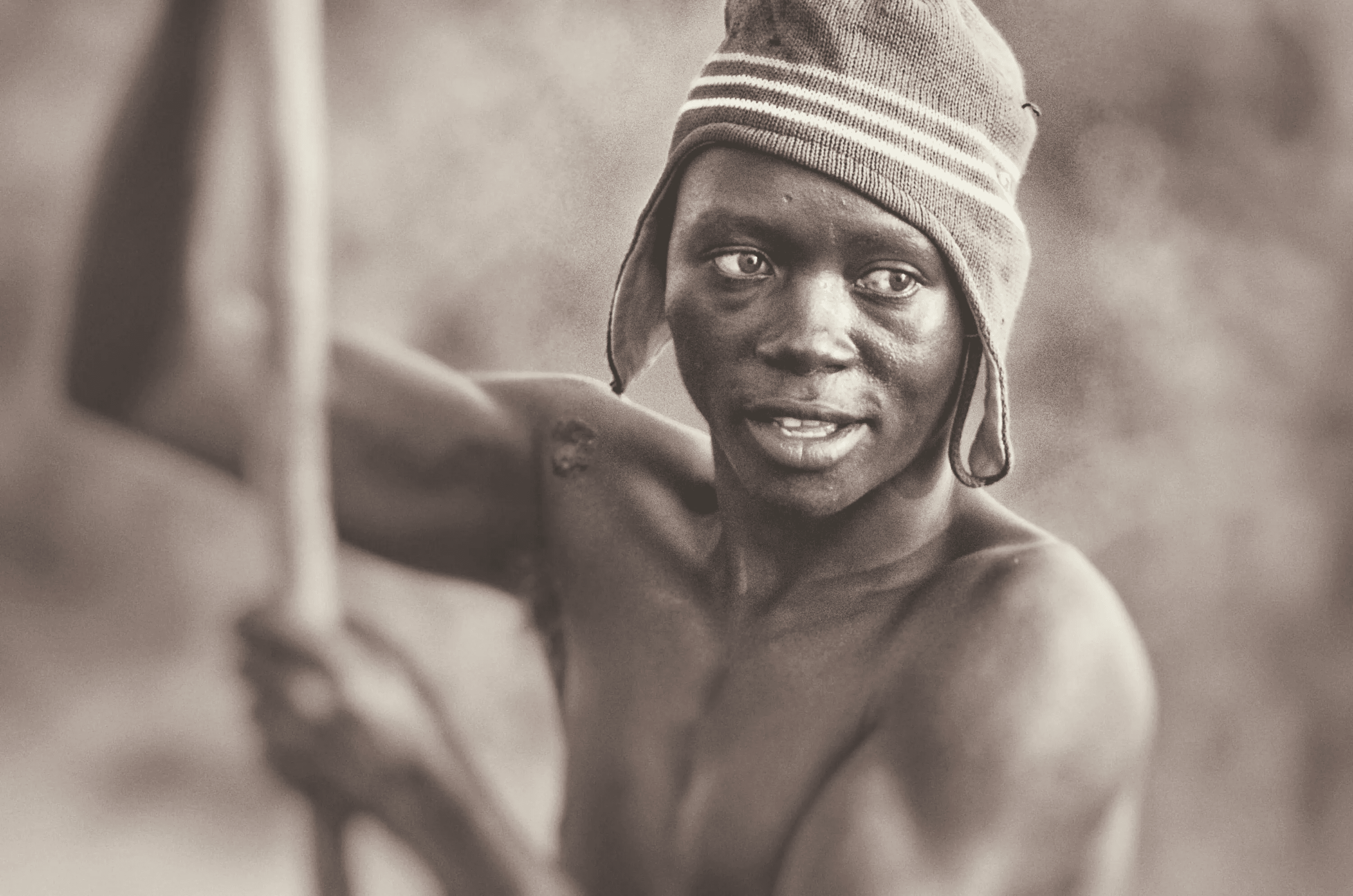 A close-up, sepia-toned portrait photograph of a Ugandan fisherman. He is wearing a knitted hat with stripes and is holding a long wooden pole, possibly a fishing rod or part of a boat. His expression is focused and engaged, looking slightly off-camera. The background is softly blurred, emphasizing the subject and creating an intimate and personal feel to the portrait.