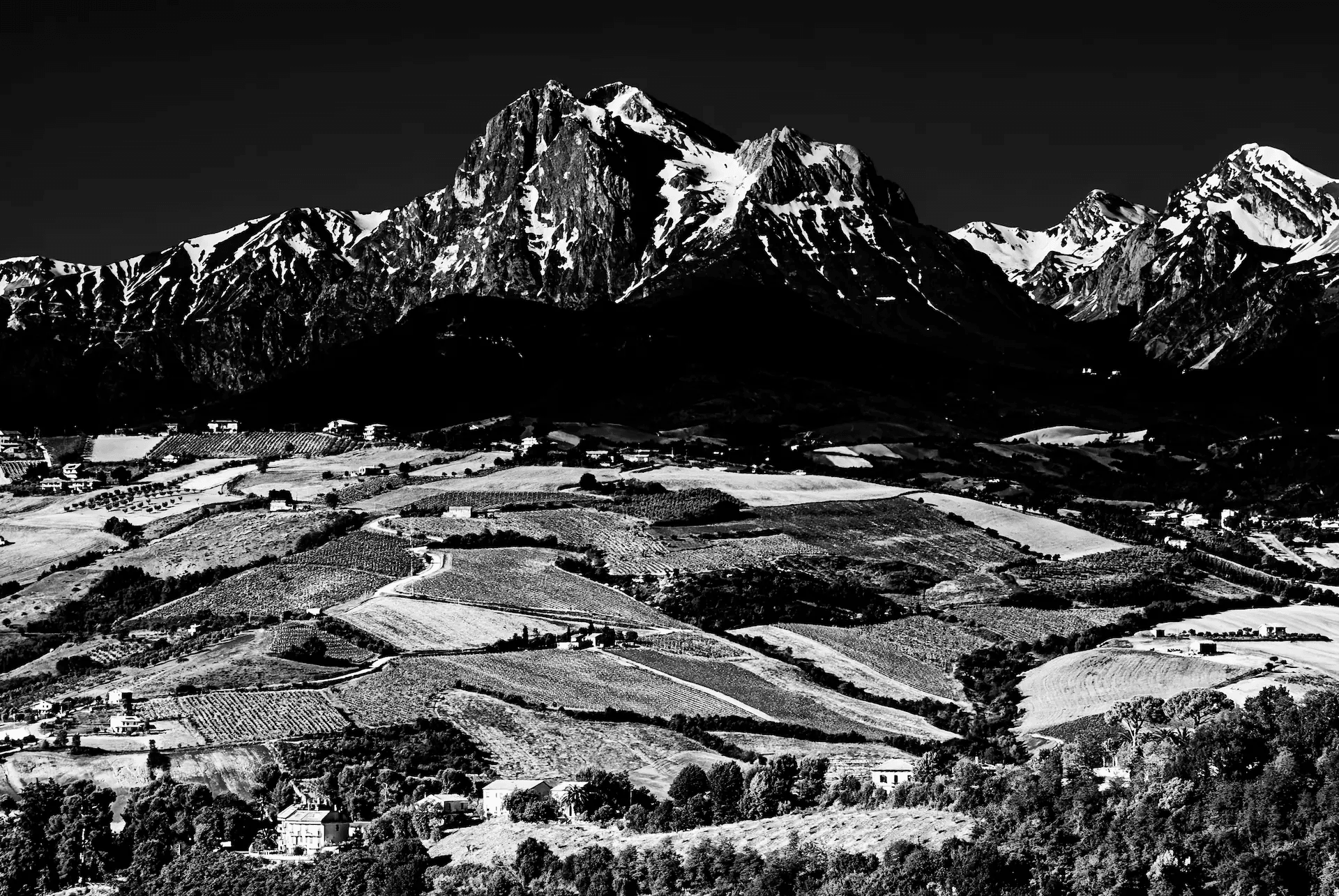 A striking black and white landscape photograph showcasing the imposing Gran Sasso mountain range in Italy. The image features rugged, snow-capped peaks dominating the skyline, their textures and contours sharply defined in monochrome. Below the mountains, rolling hills and valleys are visible, dotted with fields and small settlements. The high contrast black and white rendering enhances the dramatic quality of the scene, emphasizing the stark beauty of the Italian landscape.