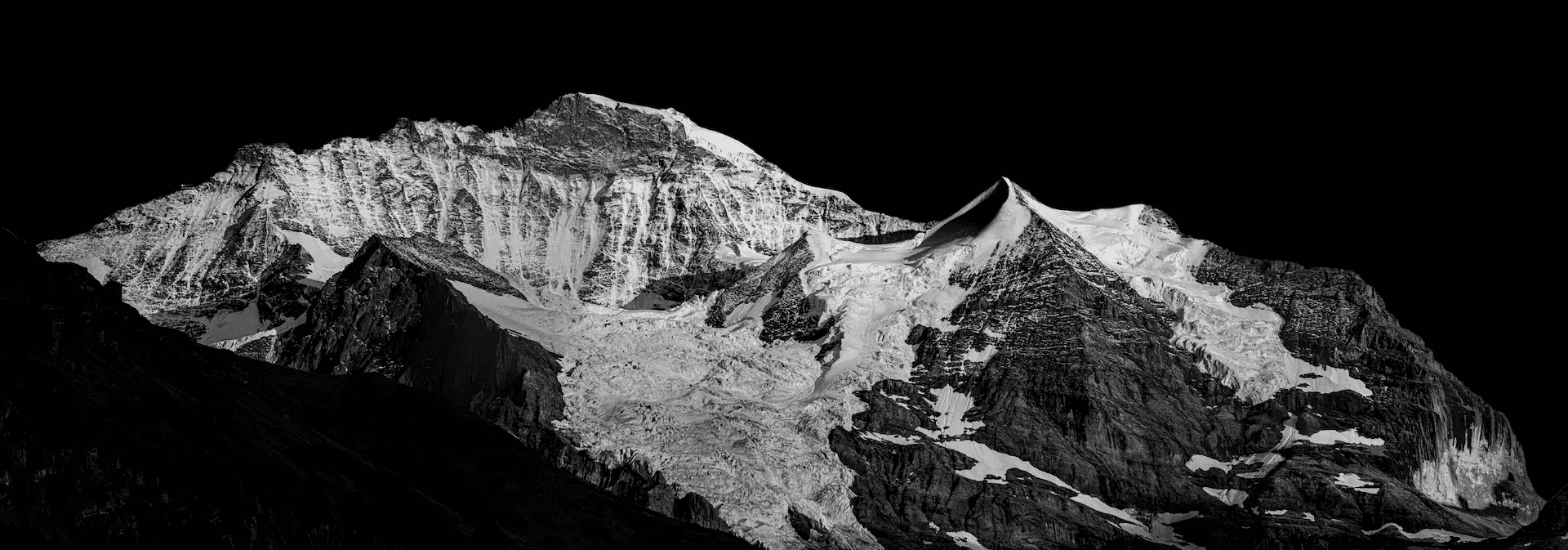 Black and white landscape photograph of the Jungfrau mountain in the Swiss Alps, viewed from Wengen. The snow-covered peak dominates the frame against a stark, dark sky.