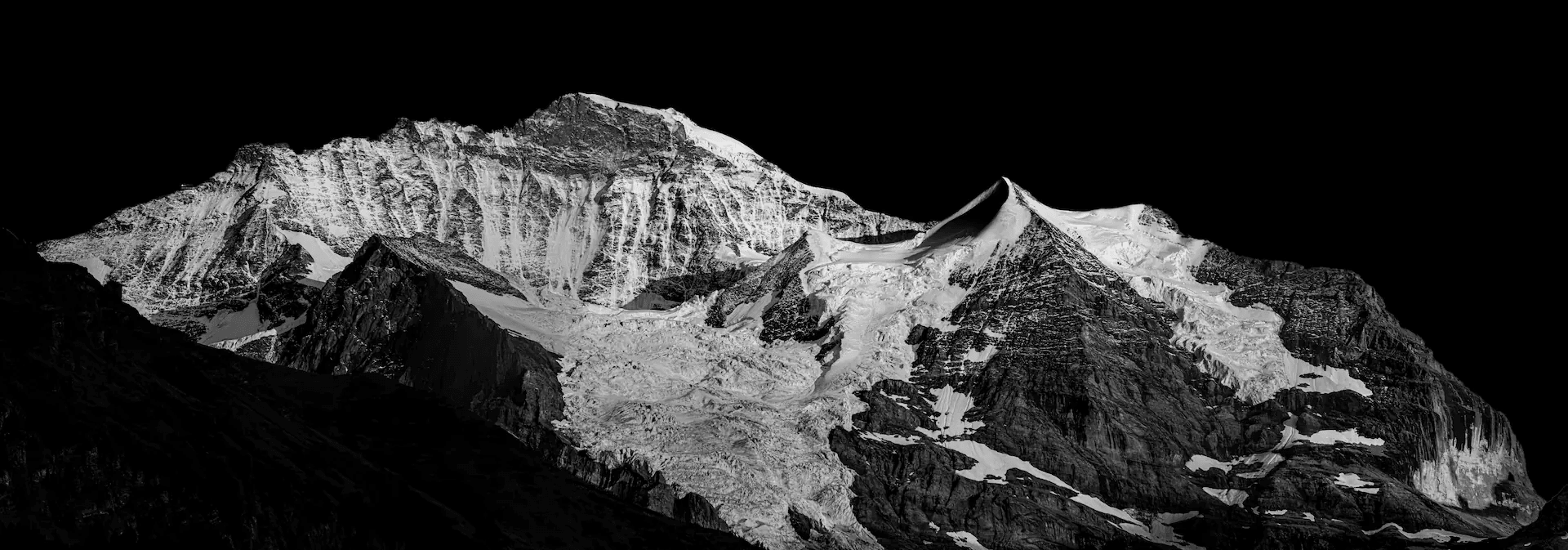 Black and white landscape photograph of the Jungfrau mountain in the Swiss Alps, viewed from Wengen. The snow-covered peak dominates the frame against a stark, dark sky. Textures of rock and snow are emphasized in monochrome. Dramatic and powerful alpine scenery.