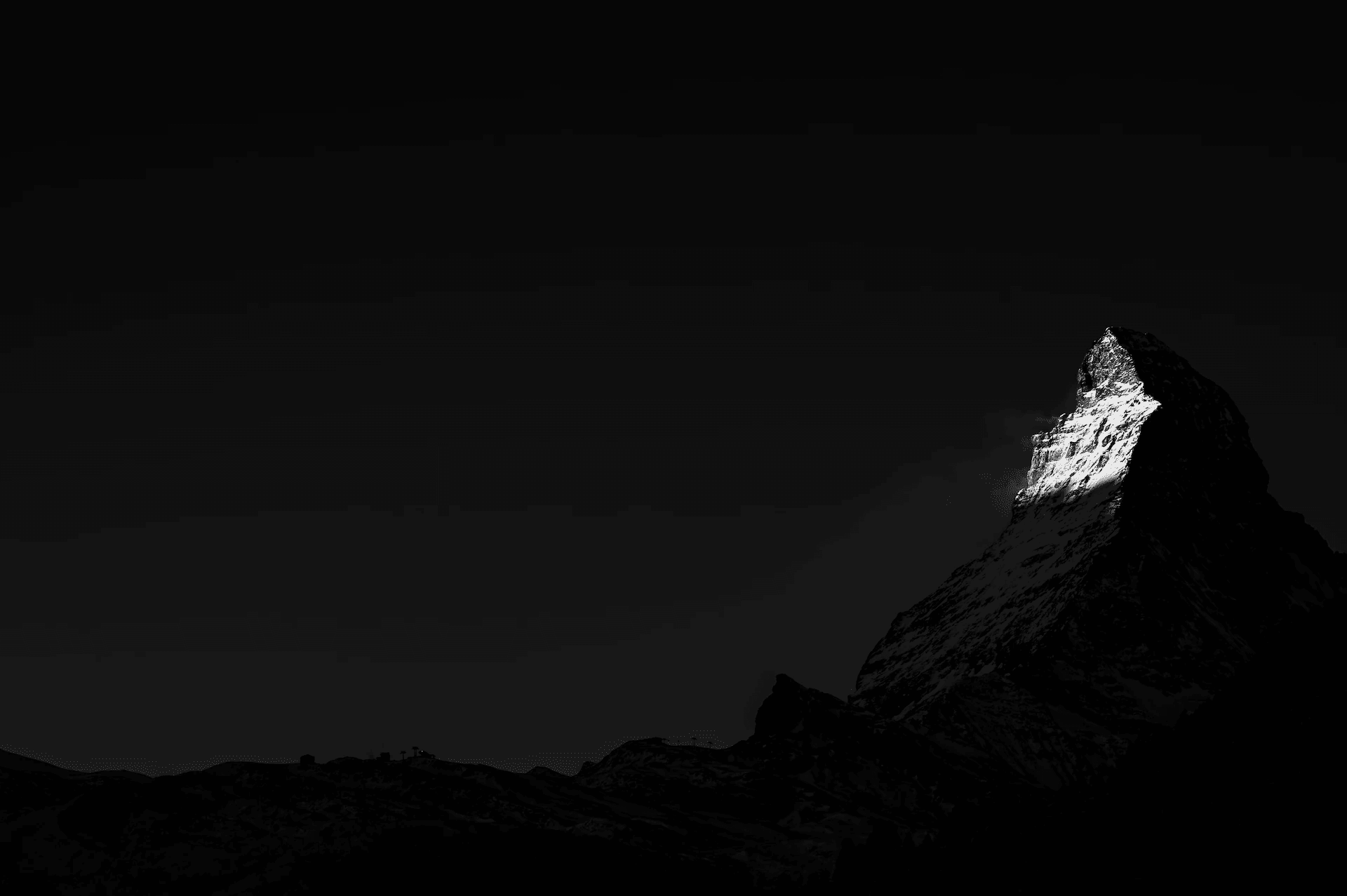 A dramatic black and white photograph of the Matterhorn mountain peak, with the first rays of morning sunlight catching the summit, while the rest of the mountain remains in shadow. The sky is still dark, suggesting the early hour, and the contrast between the brightly lit peak and the shadowed slopes is striking. The image captures the serene and powerful moment of dawn in the Swiss Alps, highlighting the iconic shape of the Matterhorn.