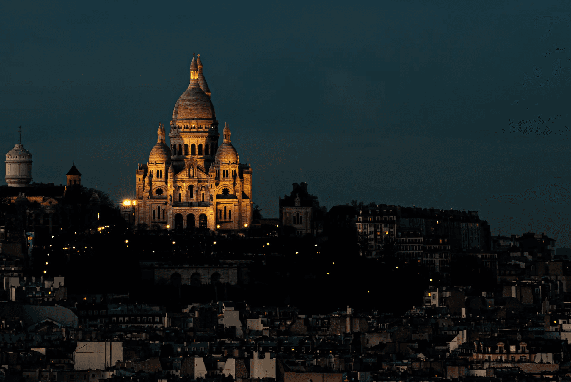 A long shot photograph capturing the Basilica of Sacré-Cœur de Montmartre in Paris, illuminated against a deep blue evening sky.