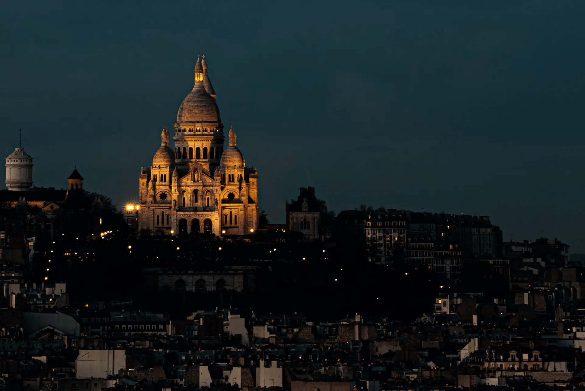 A long shot photograph capturing the Basilica of Sacré-Cœur de Montmartre in Paris, illuminated against a deep blue evening sky. The basilica, with its distinctive white domes and Romanesque-Byzantine architecture, is brightly lit, creating a warm glow that contrasts with the surrounding darkness. The city of Paris stretches out below in the foreground, with scattered lights twinkling across the urban landscape, hinting at the bustling life within the city as night descends. The atmosphere is serene and majestic, highlighting the iconic landmark's beauty and prominence.