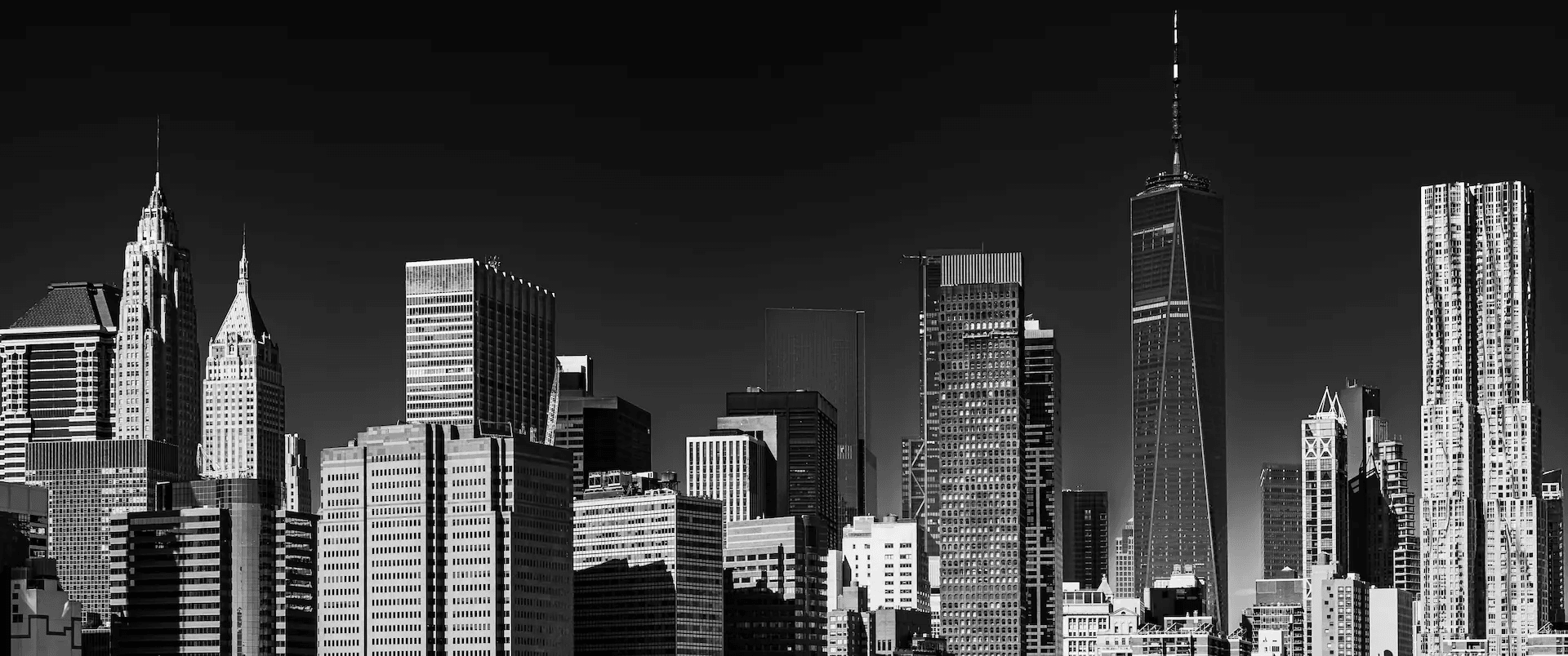 A striking black and white photograph of the New York City skyline, featuring a dense cluster of skyscrapers against a stark, clear sky. The image is rendered in high contrast, emphasizing the sharp lines and geometric forms of the architecture. Prominent buildings such as the One World Trade Center and the Chrysler Building are clearly visible, their silhouettes sharply defined. The monochromatic palette enhances the urban and architectural drama of the scene, creating a timeless and powerful representation of the iconic New York cityscape.