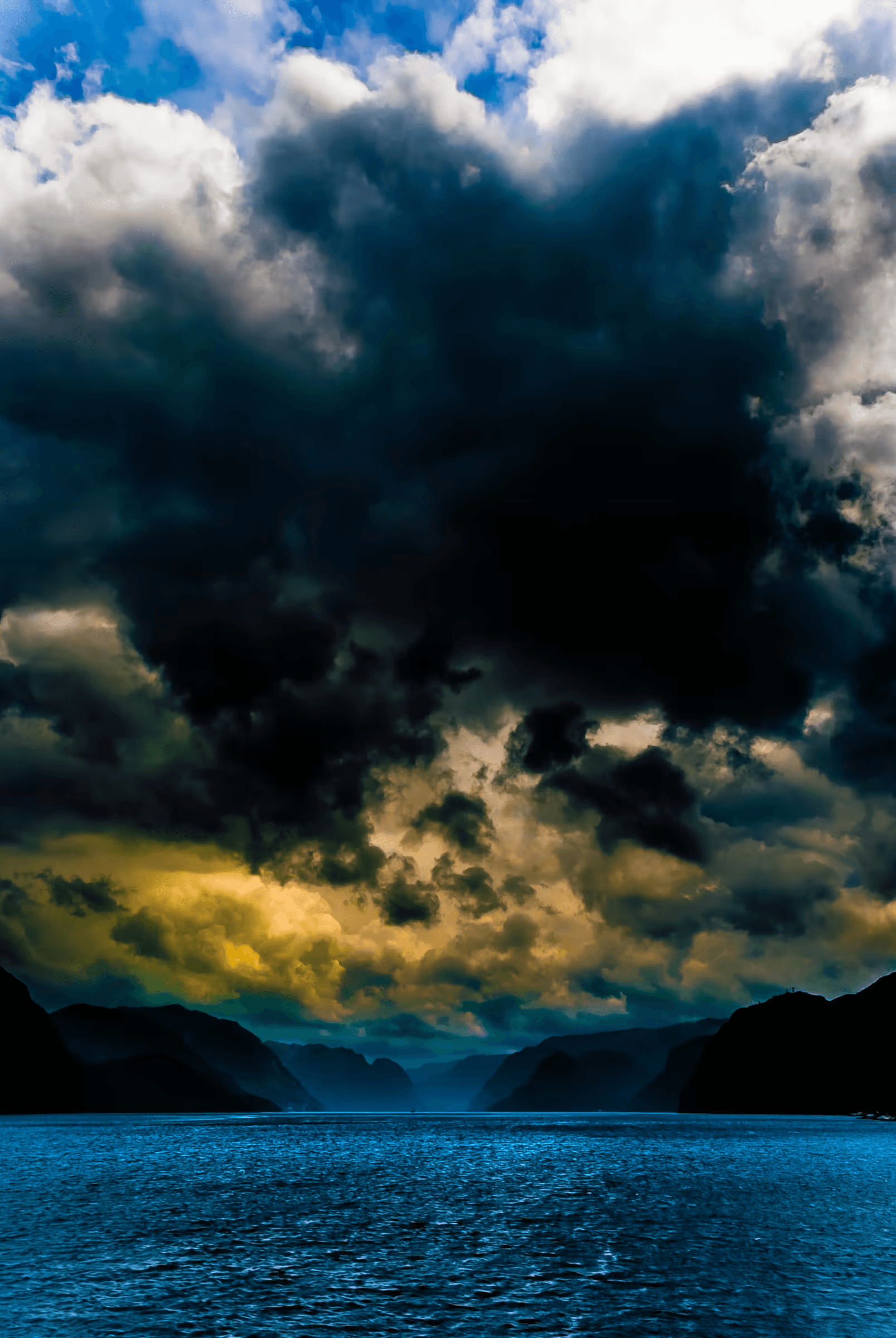 Landscape photograph of a Norwegian fjord under a dramatic, cloud-laden sky. Dark, imposing storm clouds are reflected in the tranquil fjord waters. Mountain silhouettes are visible in the distance. Evocative and atmospheric nature photography from Norway, emphasizing dramatic skies and fjord landscapes.