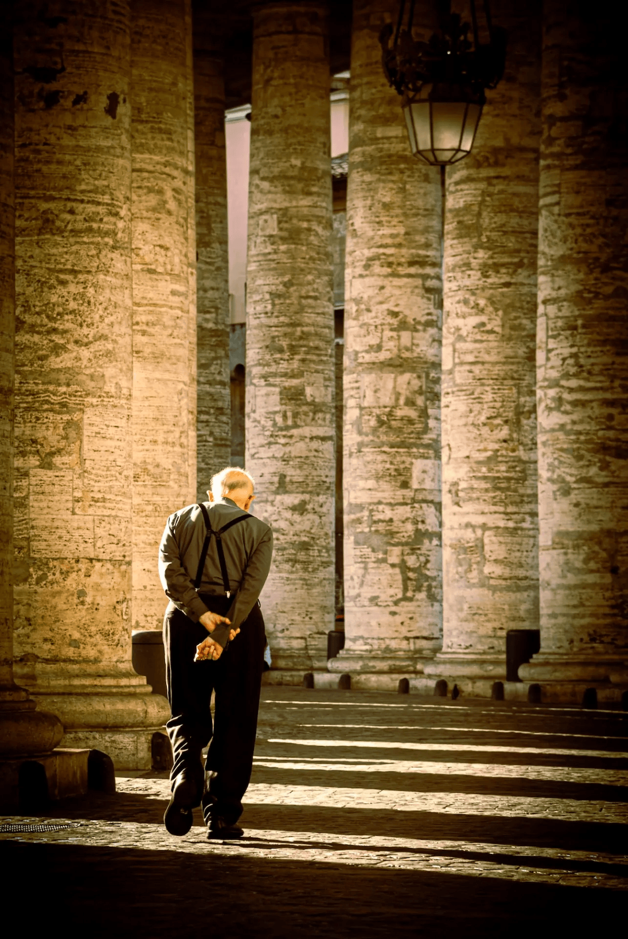 A photograph capturing an elderly priest walking away from the viewer under the grand colonnade of St. Peter's Basilica in Vatican City, Rome. The priest, dressed in dark attire with suspenders, has his hands clasped behind his back. He is walking on a cobblestone path lined by massive stone columns, casting long shadows in the warm, directional light. The architectural grandeur of the Vatican and the solitary figure of the priest create a scene of quiet reflection and urban solitude.