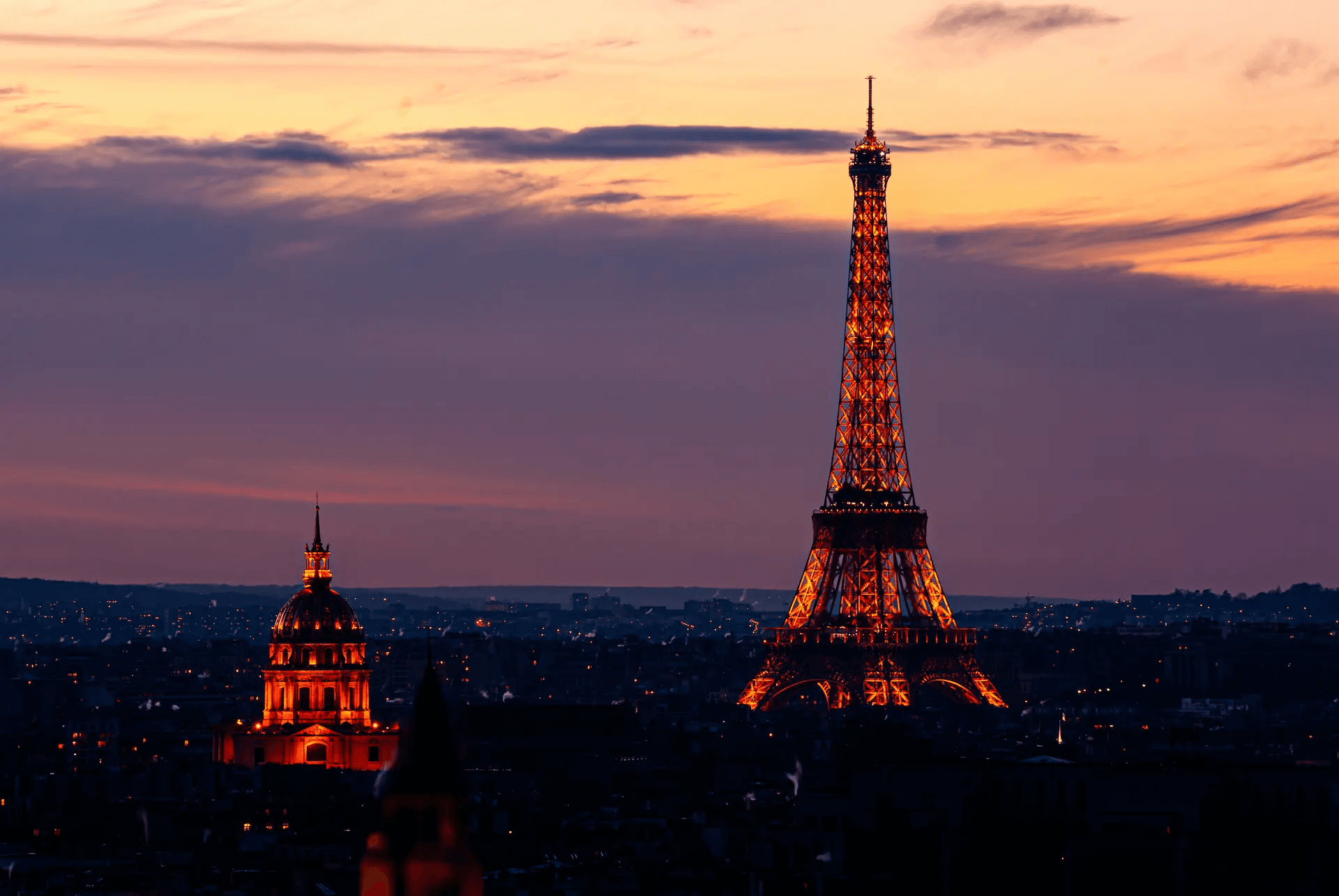 A stunning photograph of the Eiffel Tower and the dome of Les Invalides in Paris, illuminated against a vibrant twilight sky. The Eiffel Tower, the tallest structure, is bathed in warm, golden light, its intricate latticework clearly visible. To the left, the golden dome of Les Invalides also glows warmly, standing out against the darkening cityscape. The sky is a gradient of deep purples and oranges, indicative of dusk, and the city lights of Paris begin to twinkle in the background, creating a magical and romantic atmosphere.