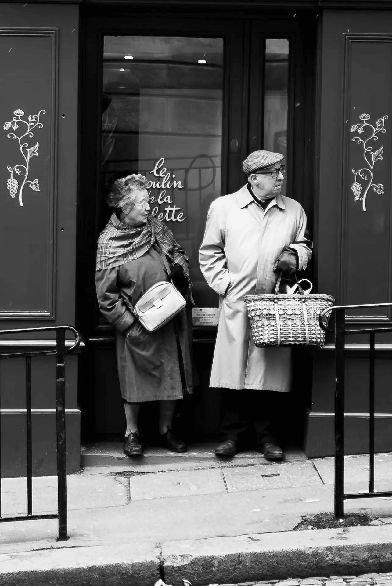 A black and white street photograph of an elderly couple standing in the doorway of a Parisian shop. The woman, on the left, is wearing a coat and scarf and carrying a handbag, looking slightly to the side with a thoughtful expression. The man, on the right, wears a cap and a trench coat, holding a woven basket and gazing off into the distance. They are positioned in a dark doorway with decorative vine-like patterns on the doorframe. The scene is captured in a candid, street photography style, suggesting a moment of quiet contemplation in the urban environment of Paris.