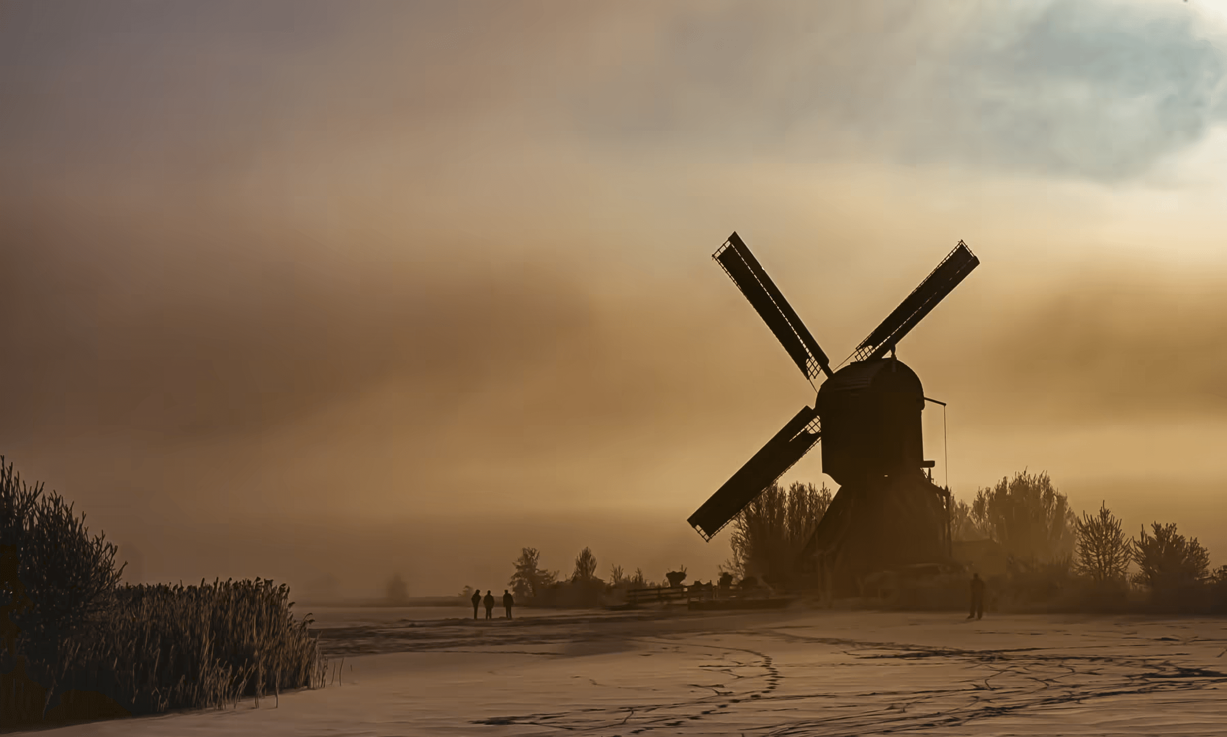 Landscape photograph of a Dutch windmill silhouetted against a misty, golden sky. The windmill's dark form stands prominently on a snowy or frosty field. Figures are visible in the distance, adding scale to the vast landscape. Golden light filters through the mist, creating a painterly effect reminiscent of Dutch Masters. Evocative winter landscape from the Netherlands.