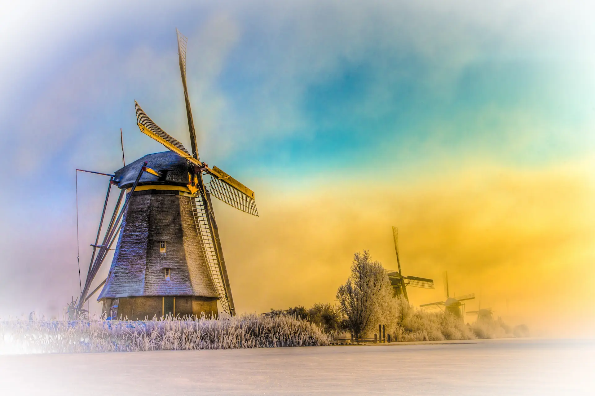 A picturesque winter landscape photograph of the windmills at Kinderdijk, Netherlands.  Several traditional windmills stand prominently against a pastel-colored sky, ranging from soft blues to warm yellows.