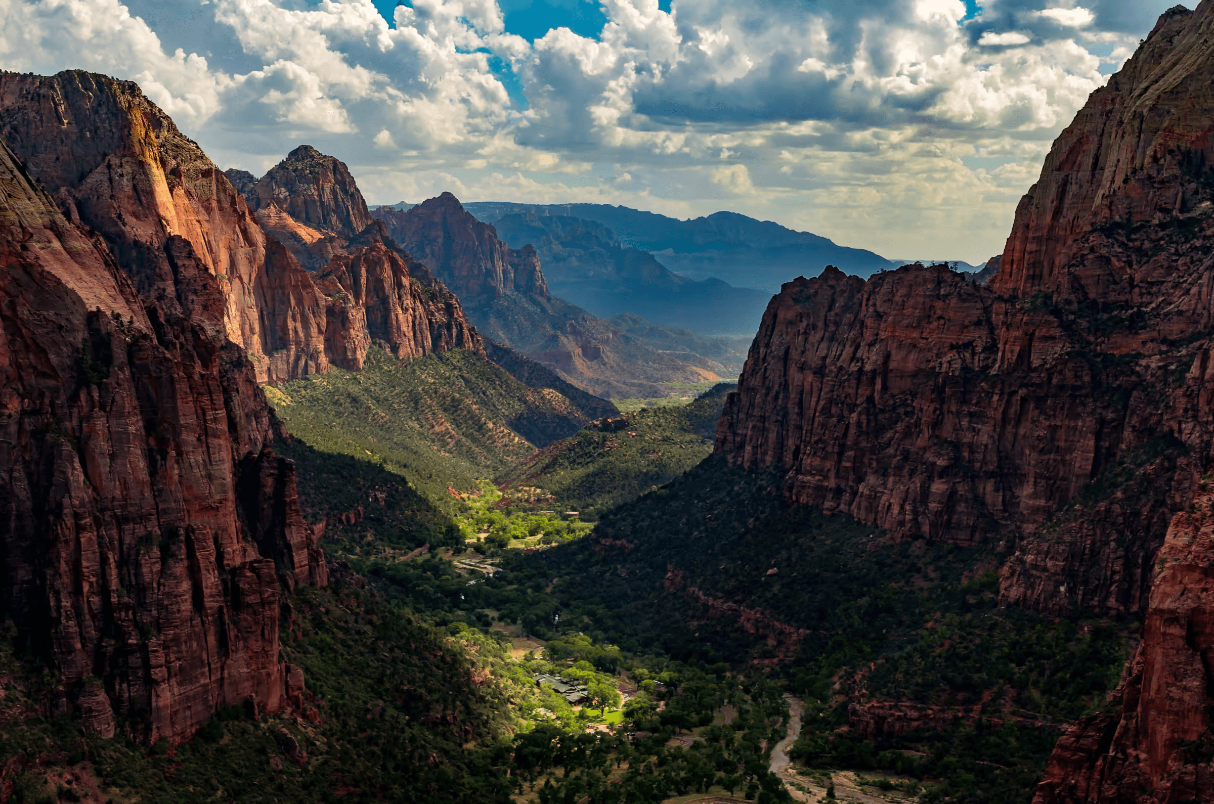 Landscape photograph capturing a panoramic vista from Angels Landing trail in Zion National Park, Utah. Expansive view of a deep canyon valley with towering red rock cliffs on either side. Lush greenery dots the valley floor, and distant mountains fade into the horizon under a sky filled with dramatic white and grey clouds. Daytime nature scene showcasing the grandeur of Zion National Park.