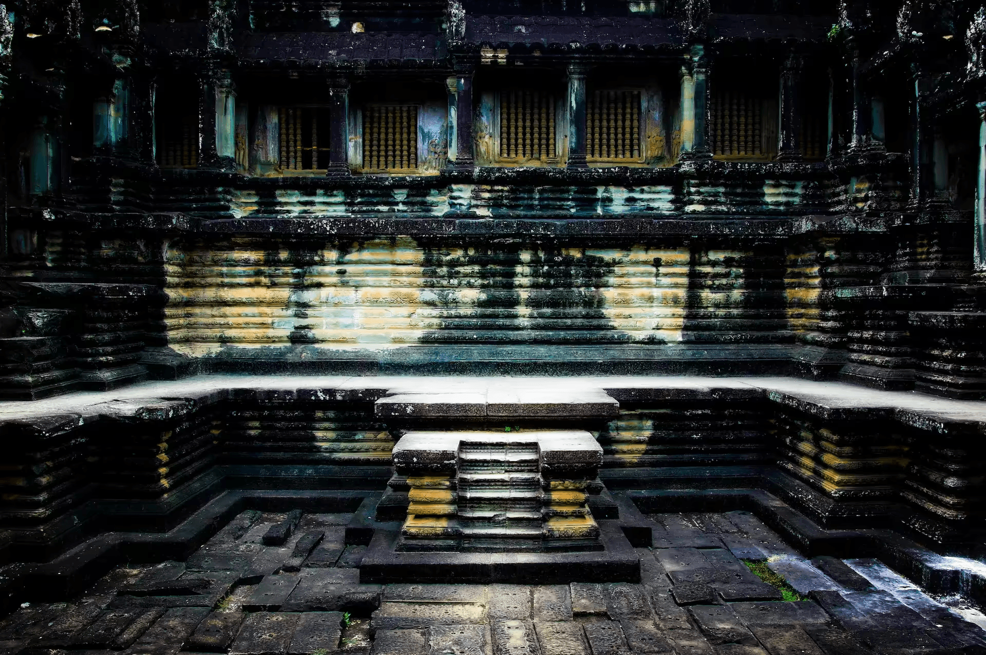 A detailed photograph showcasing the interior stone architecture of Angkor Wat temple in Cambodia. The image focuses on a section of the temple with layered stone walls, columns, and window openings with decorative latticework. The ancient stone surfaces are weathered and textured, showing signs of age and history. The lighting is dramatic, casting shadows and highlighting the intricate carvings and architectural details of this UNESCO World Heritage site. The scene evokes a sense of ancient grandeur and the enduring legacy of Khmer architecture.