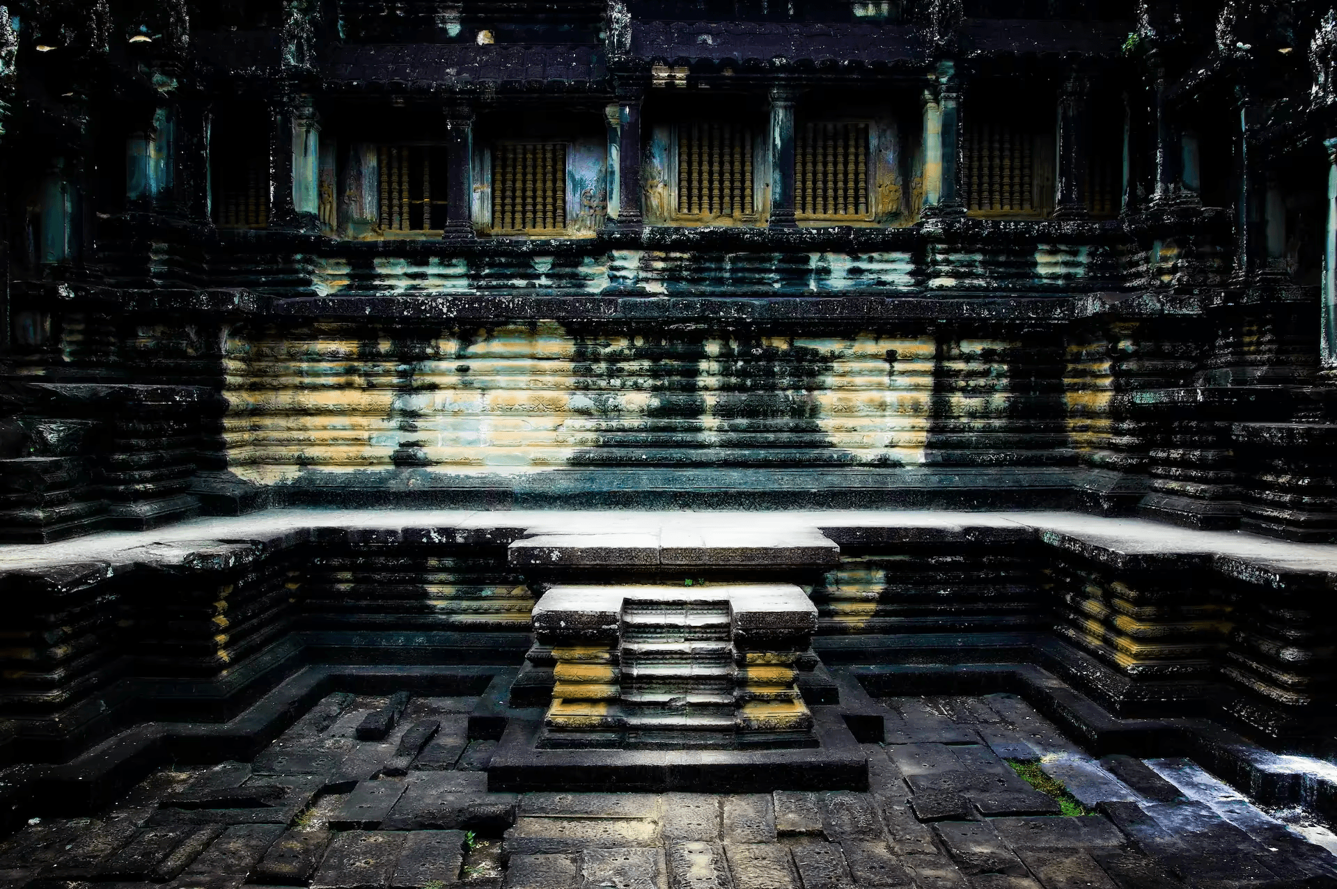 A detailed photograph showcasing the interior stone architecture of Angkor Wat temple in Cambodia. The image focuses on a section of the temple with layered stone walls, columns, and window openings with decorative latticework.
