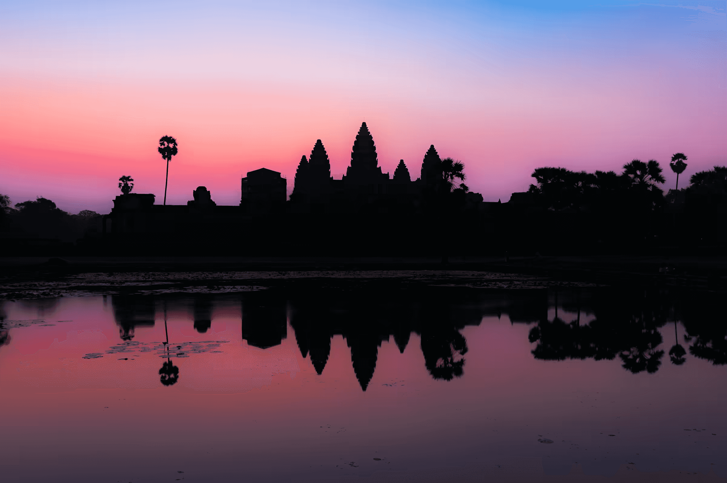 Dawn photograph of Angkor Wat temple complex silhouetted against a gradient sky, with the ancient spires and palm trees perfectly reflected in still water.