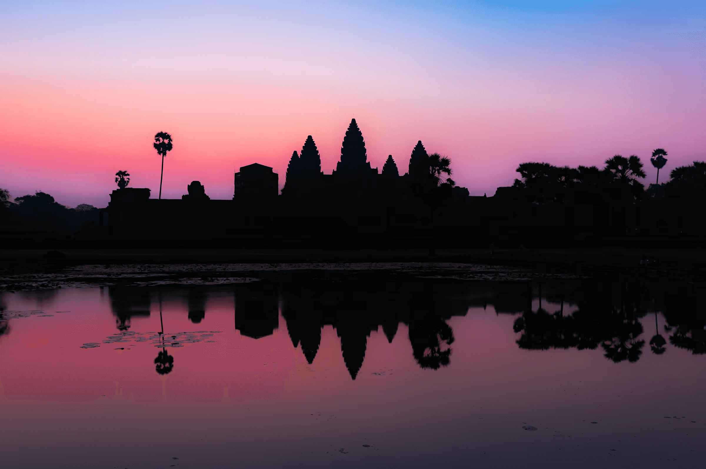Dawn photograph of Angkor Wat temple complex silhouetted against a gradient sky, with the ancient spires and palm trees perfectly reflected in still water.