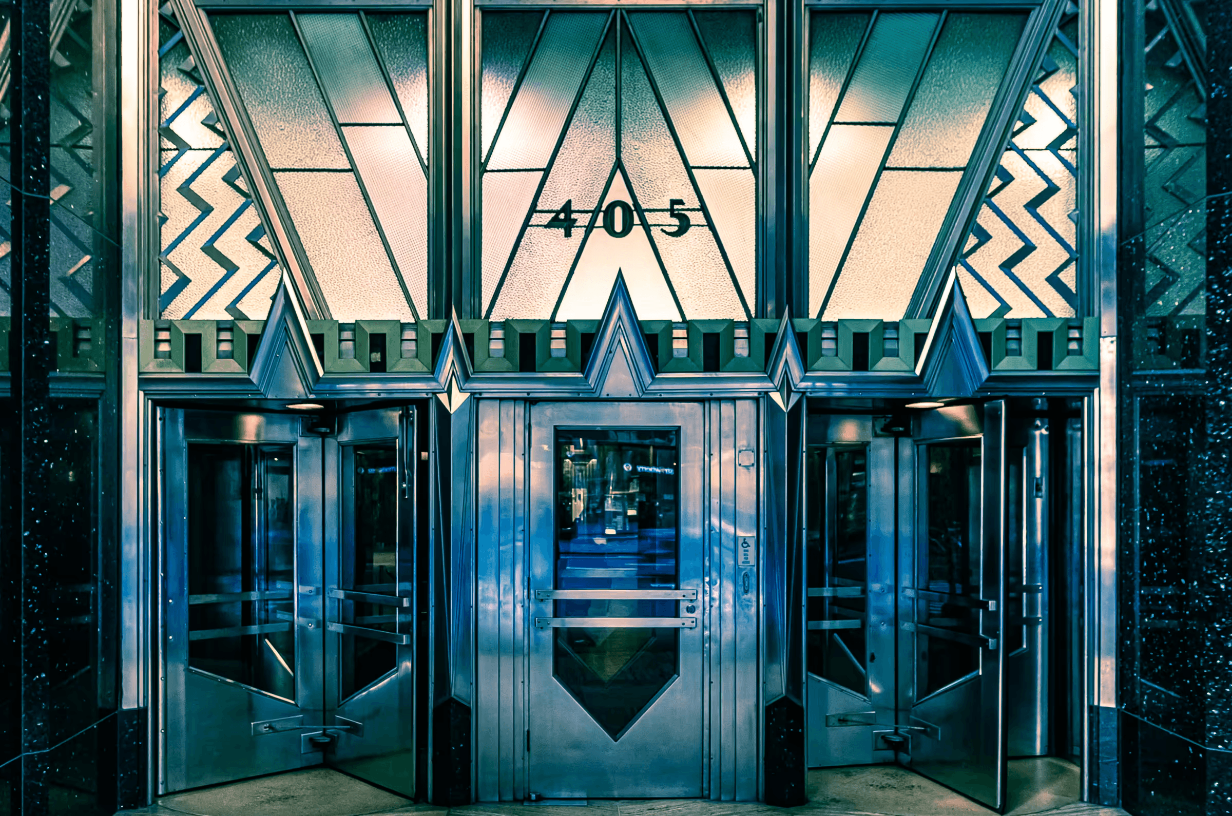 A detailed photograph of the Art Deco-style entrance doors of the Chrysler Building in Manhattan, New York City. The image highlights the intricate geometric patterns, polished metal surfaces, and stylized ornamentation characteristic of Art Deco architecture, with a hint of teal in the glass and metalwork.