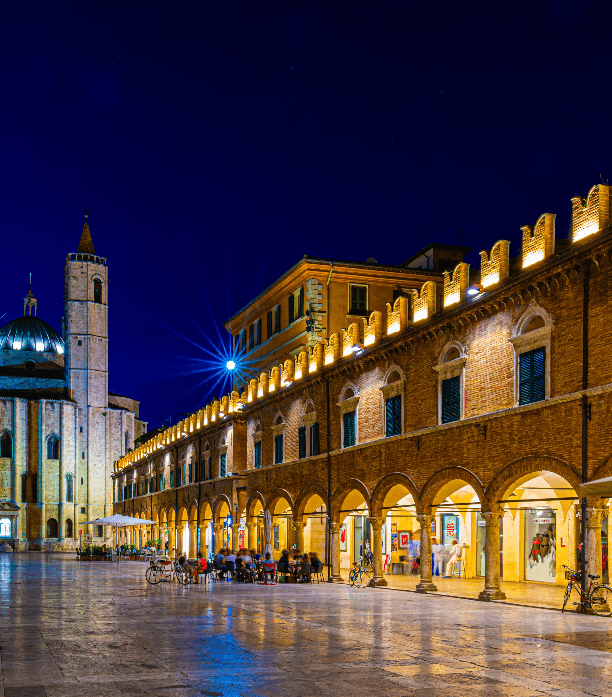 A captivating night photograph of Piazza del Popolo in Ascoli Piceno, Italy, showcasing illuminated Renaissance buildings with elegant colonnades. The warm glow of lights highlights the historic architecture and the cobblestone pavement of the piazza under a dark evening sky. A church tower is subtly visible in the background.