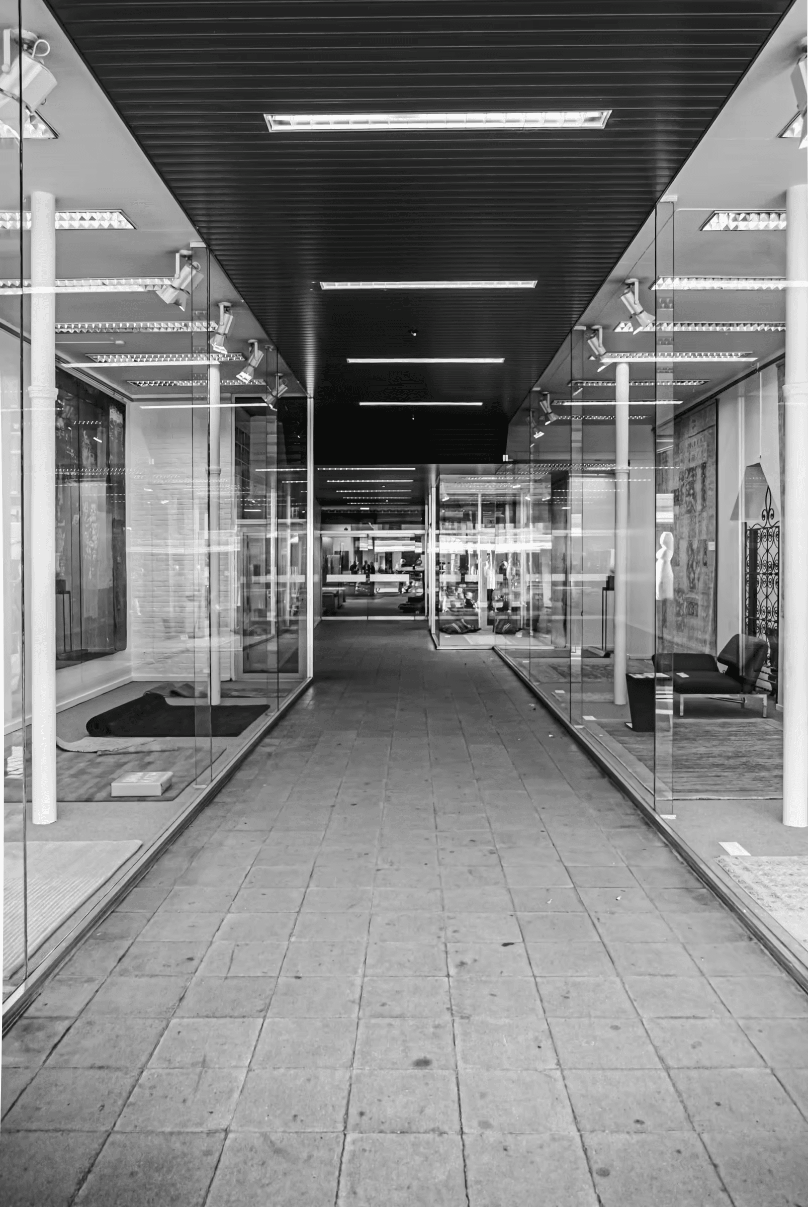 Black and white photograph of a modern city corridor. Strong geometric lines are formed by the architecture, including glass walls and a tiled floor.