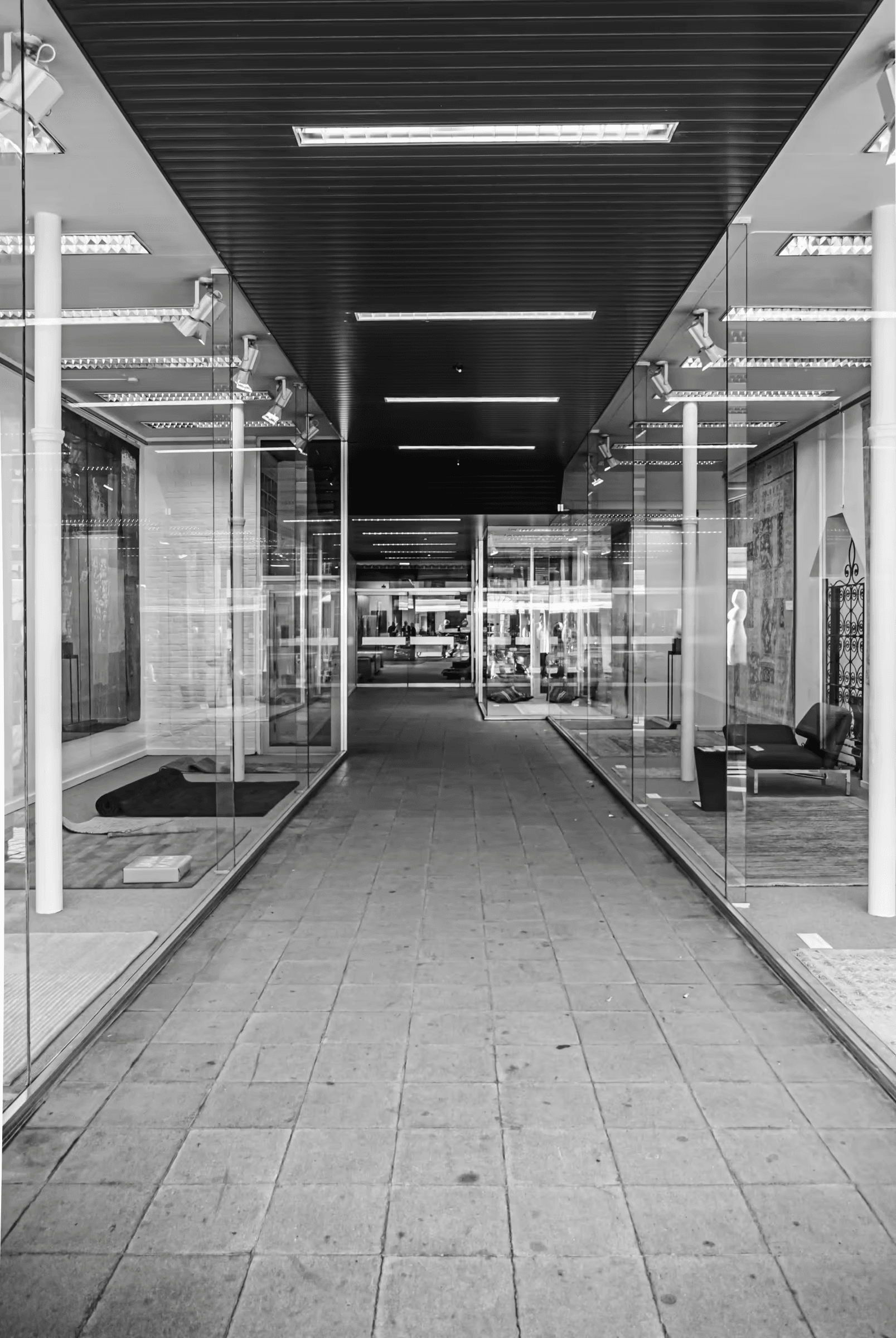 Black and white photograph of a modern city corridor. Strong geometric lines are formed by the architecture, including glass walls and a tiled floor. The perspective leads the eye down the corridor. Urban architecture, geometric, monochrome, city scene, corridor, walkway.