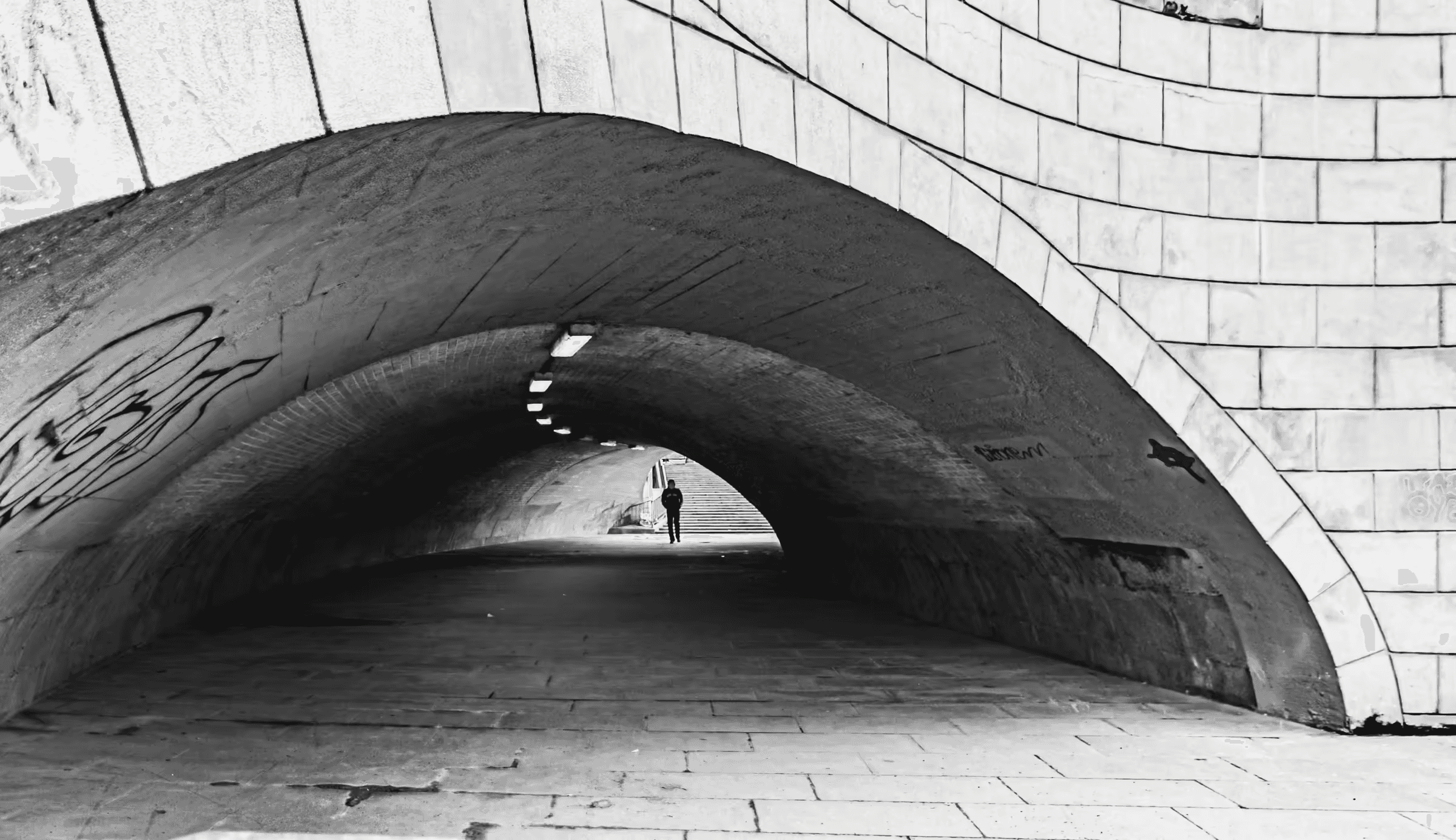 Black and white photograph of an arched city tunnel. The tunnel's brick or stone construction is visible in monochrome.