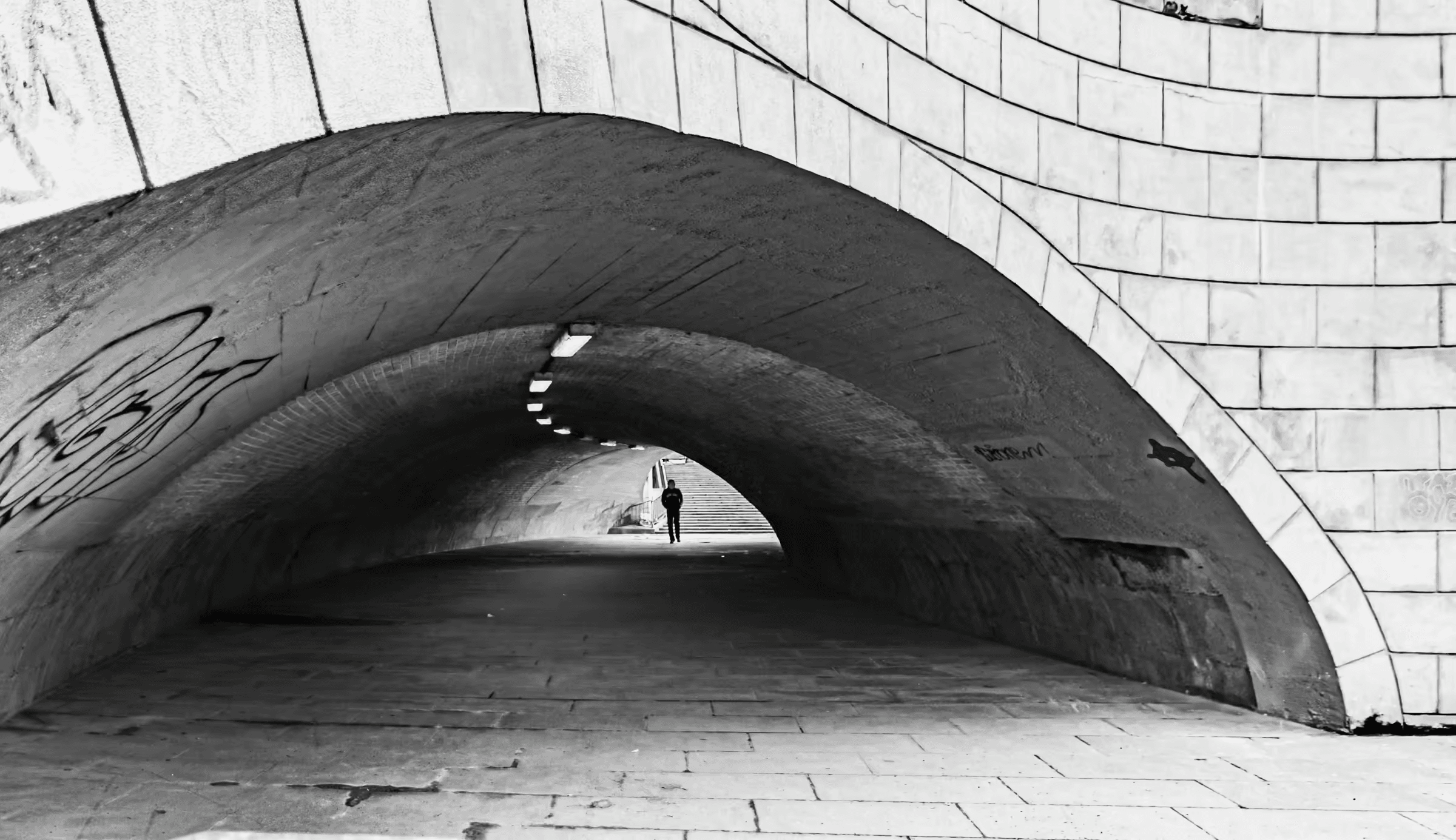 Black and white photograph of an arched city tunnel. The tunnel's brick or stone construction is visible in monochrome. A lone person is seen walking into the light at the far end of the tunnel, creating a sense of depth and perspective. Urban architecture in monochrome, tunnel, passageway, city scene.