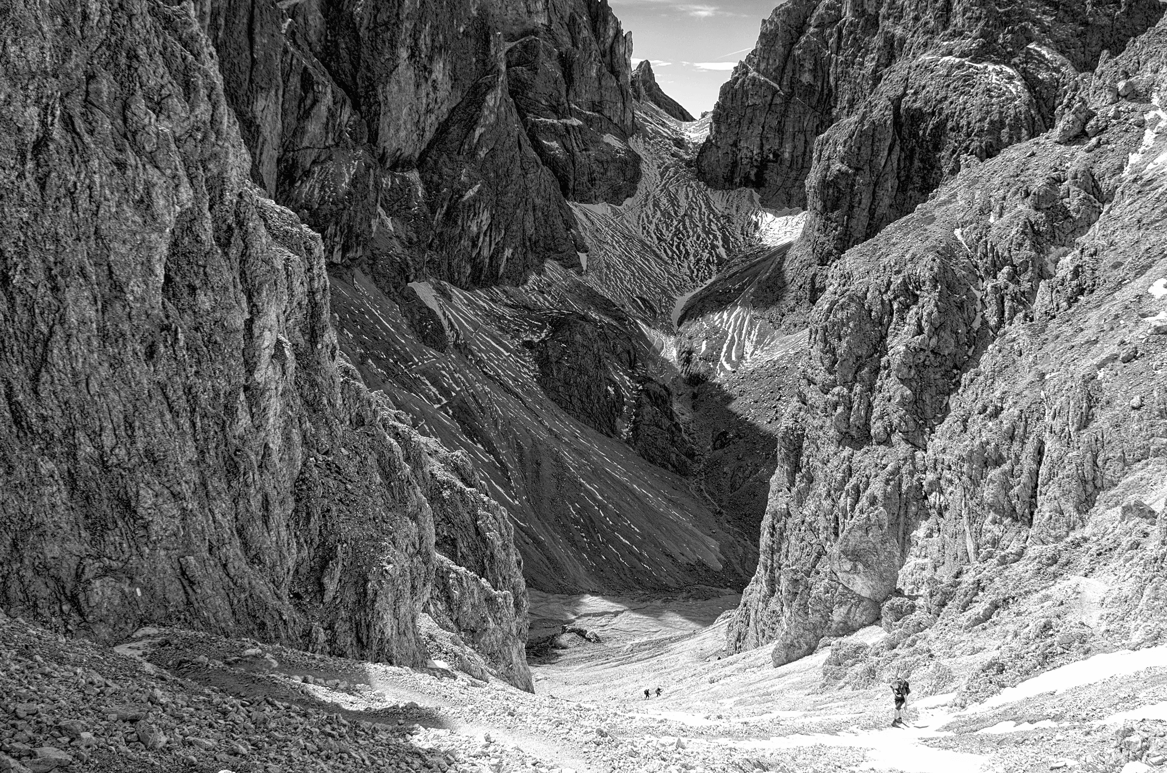 Black and white photograph looking down into a vast, rocky alpine valley near Grasleitenhütte in the Dolomites, Italy, with tiny hikers visible on a snowfield below.