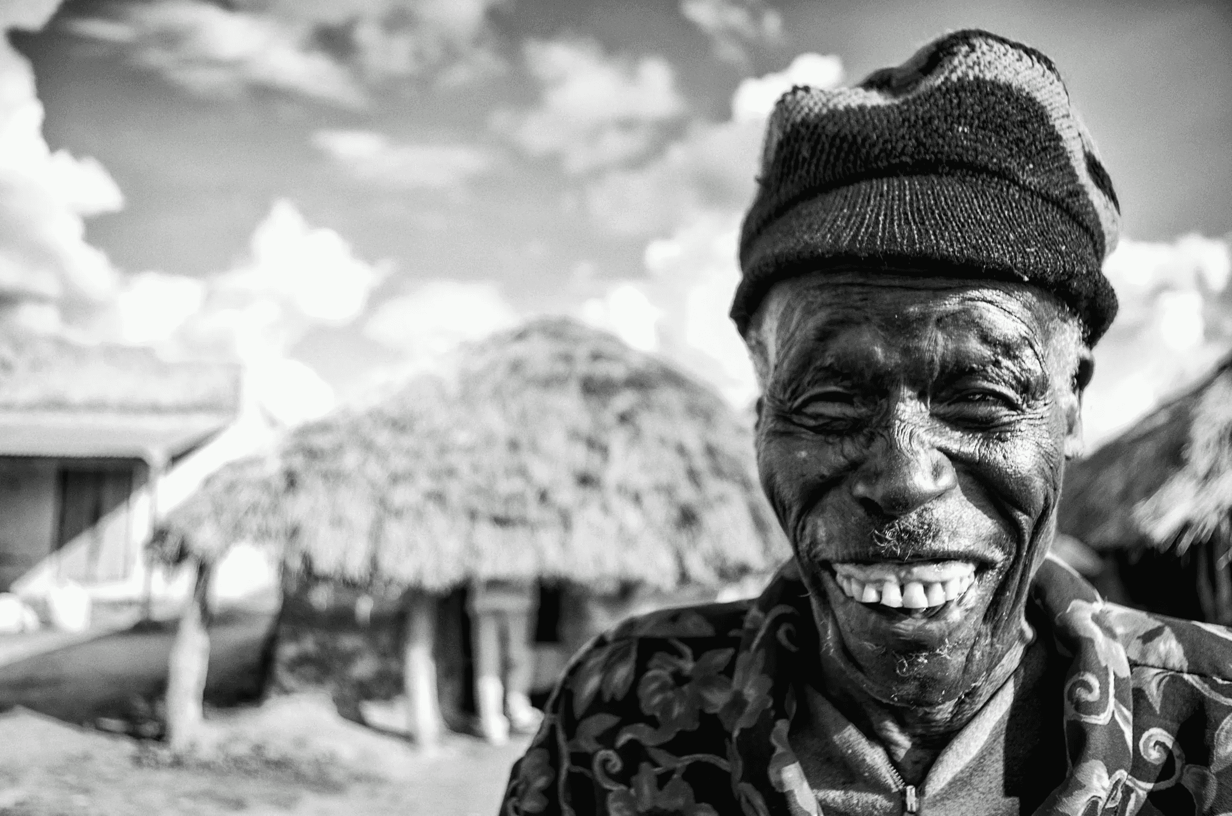 Black and white portrait of a smiling elderly man wearing a knit cap and patterned shirt, outdoors in Amuria, Uganda, with traditional thatched-roof huts blurred in the background..