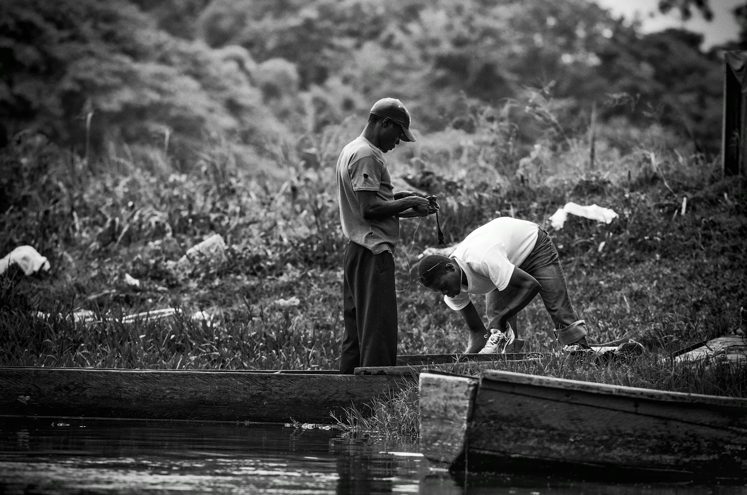 Black and white photograph showing two men working on or near simple boats/canoes on a riverbank, possibly preparing fishing gear.