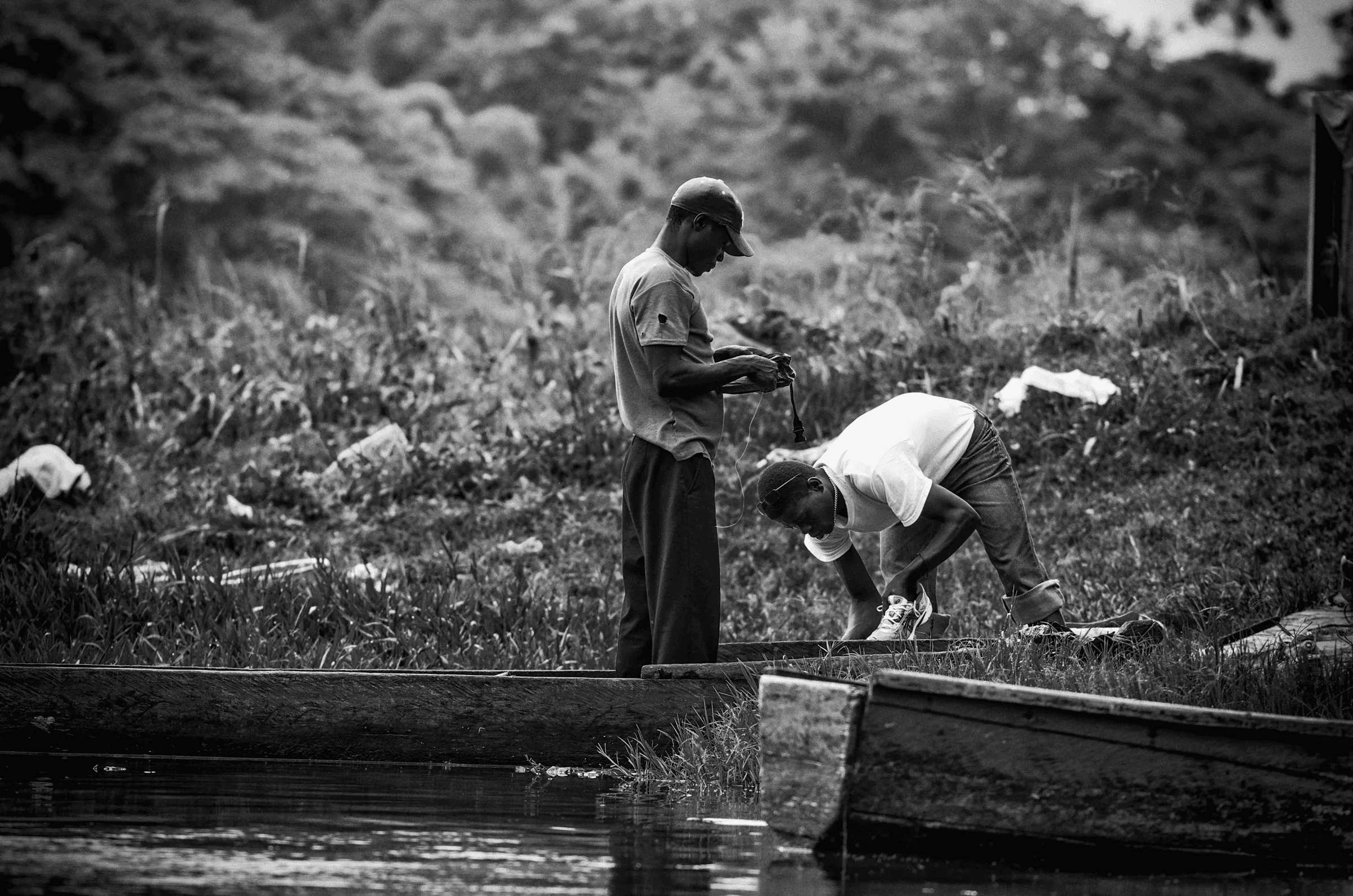 Black and white photograph showing two men working on or near simple boats/canoes on a riverbank, possibly preparing fishing gear.
