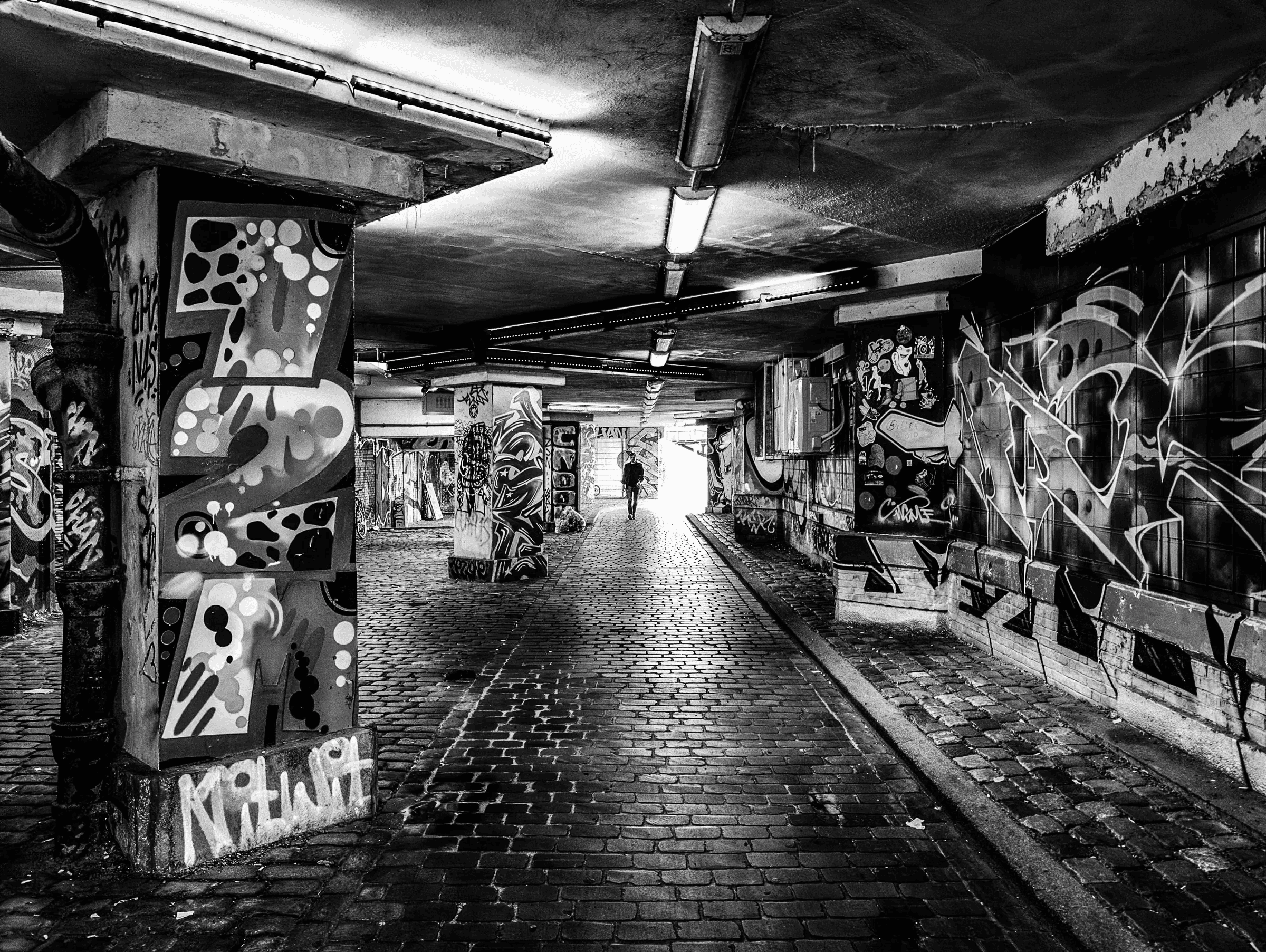 Graffiti Passage Black and white photograph of a person walking through a graffiti-covered urban underpass with strong overhead lighting and wet cobblestones.