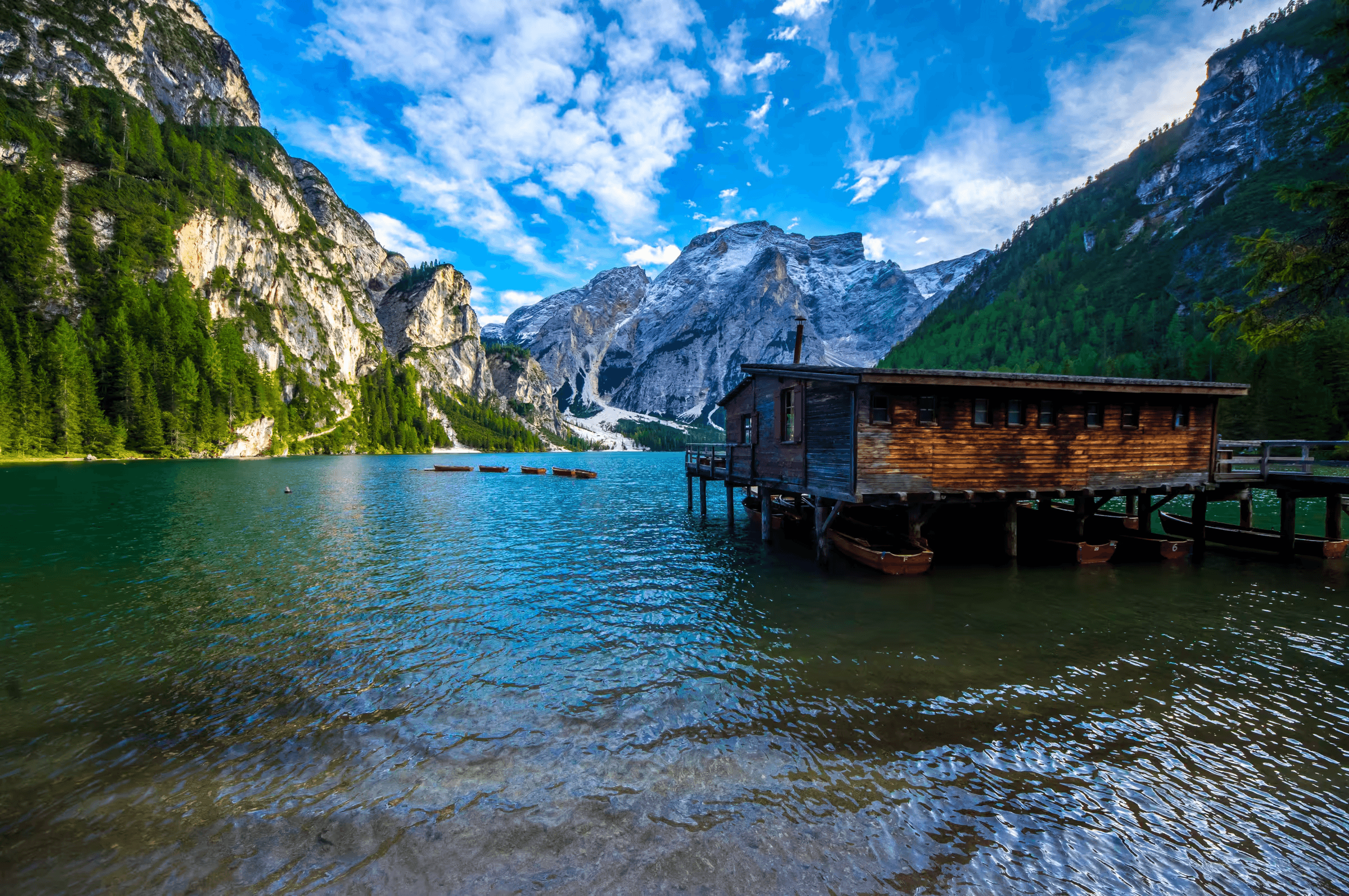 Landscape photograph of Lago di Braies (Pragser Wildsee) in the Dolomites, Italy. A wooden boathouse sits on the edge of the turquoise lake.  Several small boats are docked nearby.  Towering mountains, partially covered in forest, rise dramatically in the background under a blue sky with white, puffy clouds.