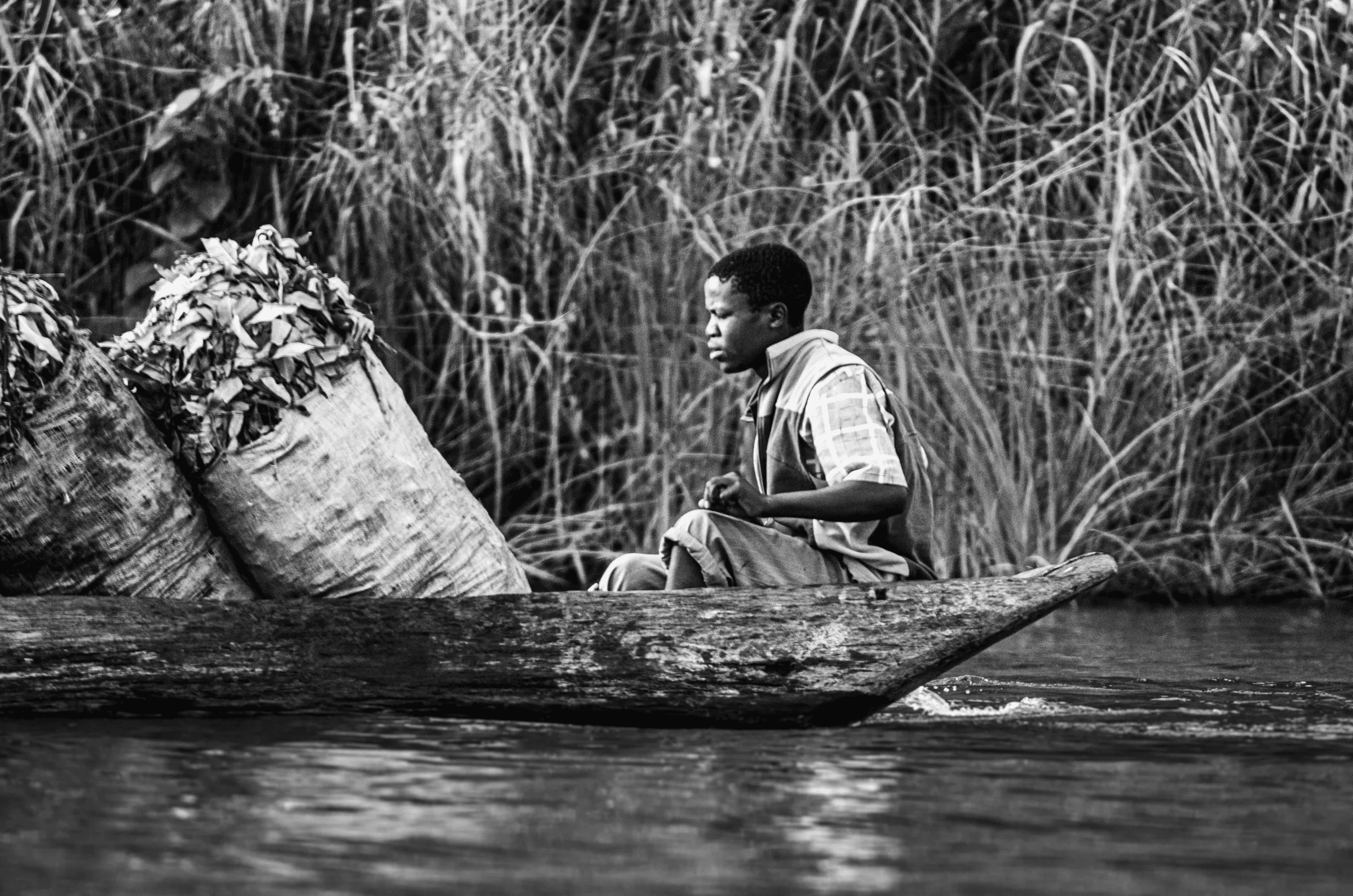 Black and white profile view of a person sitting in a dugout canoe carrying large bundles of tea on Lake Bunyonyi, Uganda.