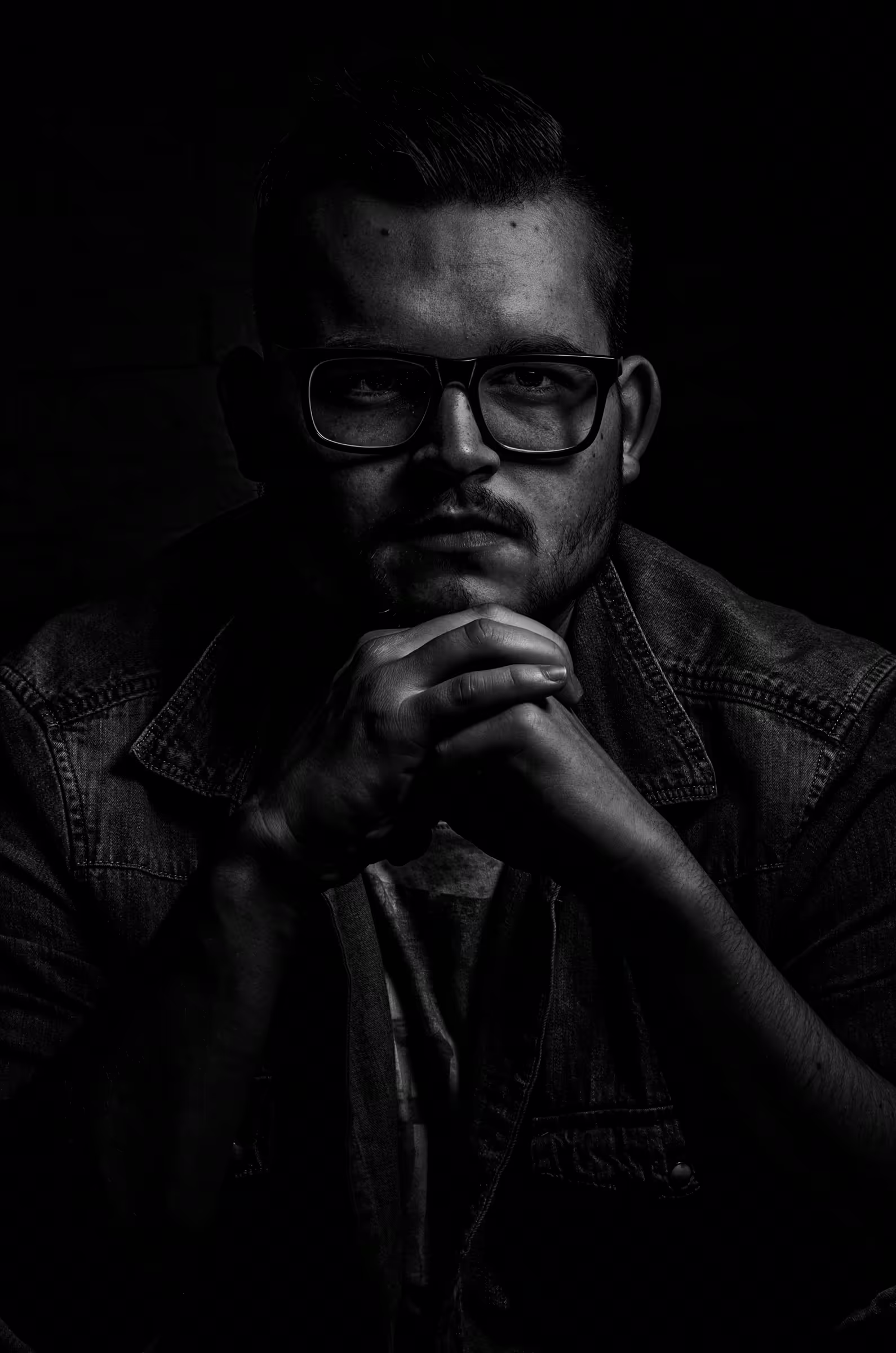 Low-key black and white portrait photograph of a man wearing glasses. The subject is positioned to the right side of the frame, his face partially illuminated, with the rest of the image in deep shadow.