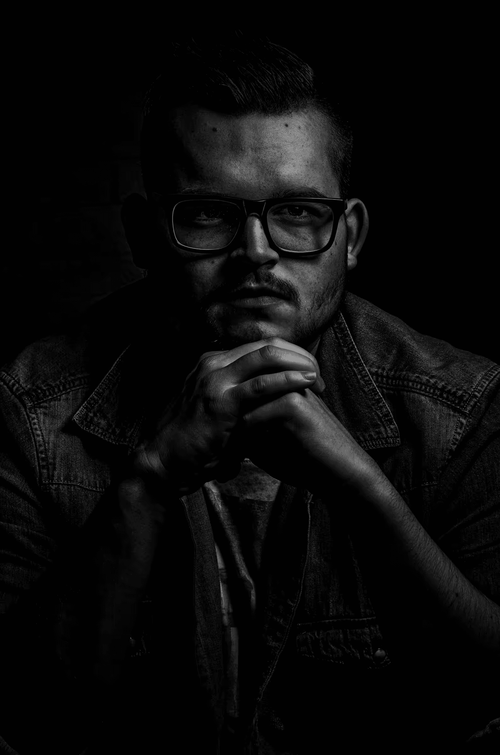 Low-key black and white portrait photograph of a man wearing glasses. The subject is positioned to the right side of the frame, his face partially illuminated, with the rest of the image in deep shadow.