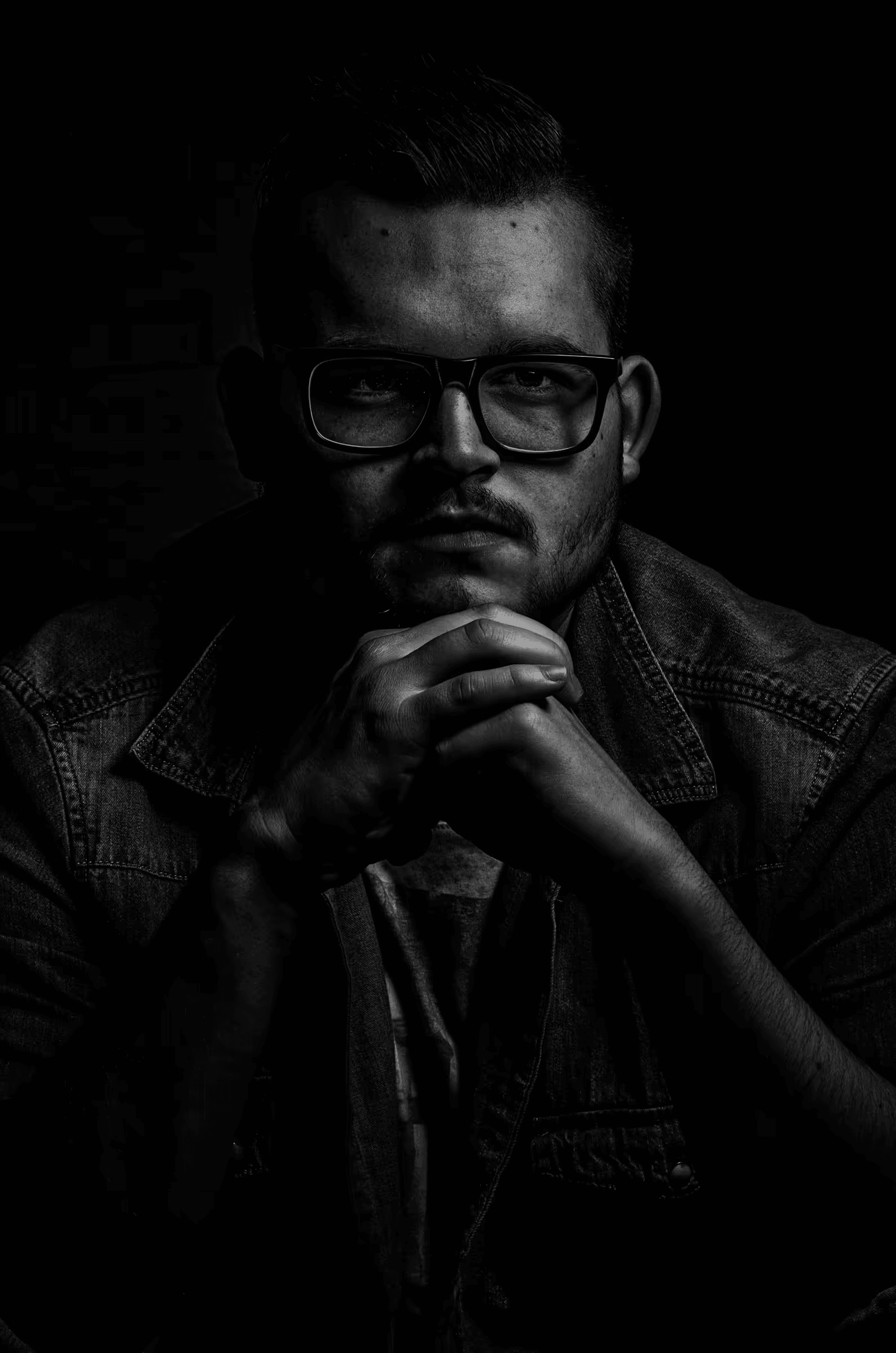 Low-key black and white portrait photograph of a man wearing glasses. The subject is positioned to the right side of the frame, his face partially illuminated, with the rest of the image in deep shadow. He is gazing slightly downwards with a pensive expression, his hands clasped in front of him. Intimate studio portrait in monochrome. 