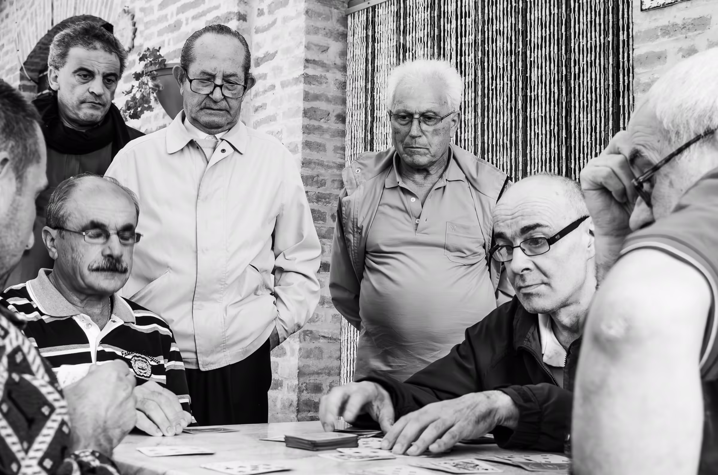 Black and white photograph of a group of older men playing cards. The men are intently focused on their cards and the game, their expressions serious and engaged.