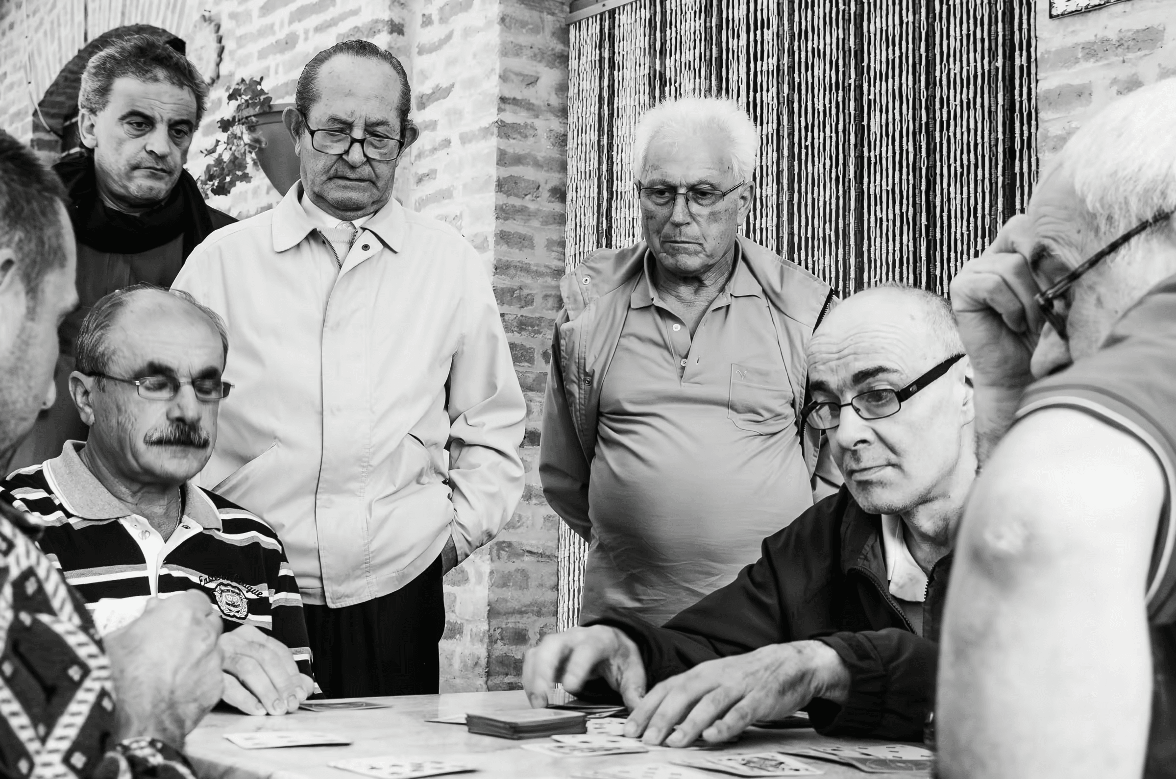 Black and white photograph of a group of older men playing cards. The men are intently focused on their cards and the game, their expressions serious and engaged.  Monochrome life photography, card game, men, gathering, focused, black and white portrait, friendship.