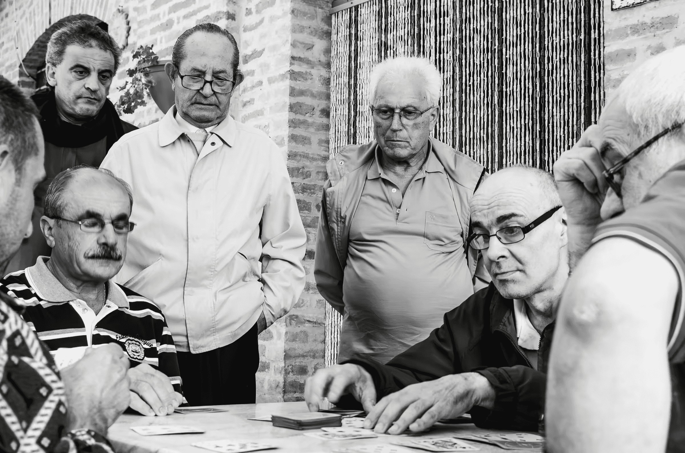 Black and white photograph of a group of older men playing cards. The men are intently focused on their cards and the game, their expressions serious and engaged.