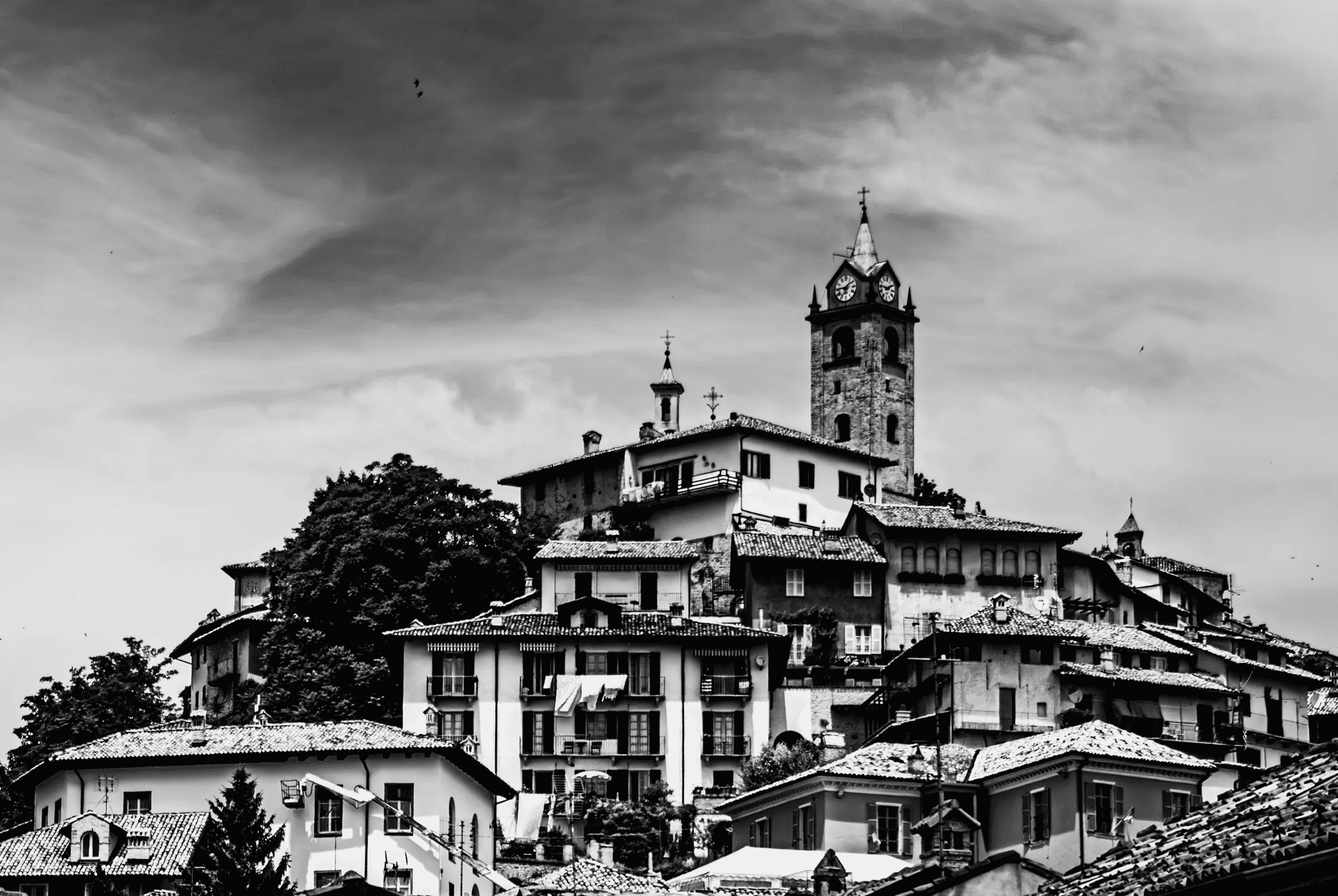 Black and white photograph looking up at the hilltop town of Monforte d'Alba, Piedmont, Italy, showing buildings clustered around a prominent clock tower under a dramatic sky.