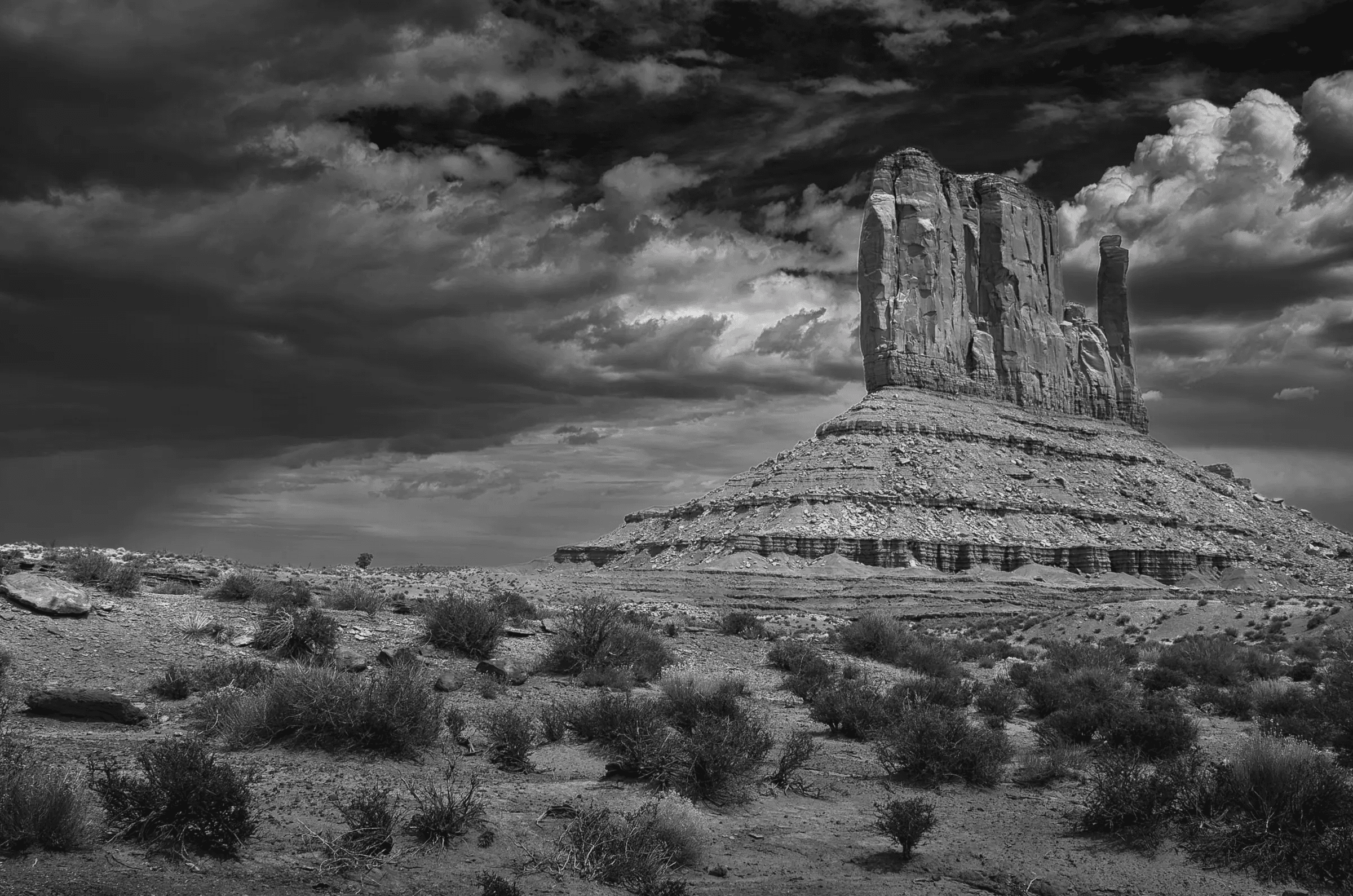 Monolith Under Weather Black and white photograph of a massive desert butte beneath dramatic, textured storm clouds, with sparse scrub in the foreground.