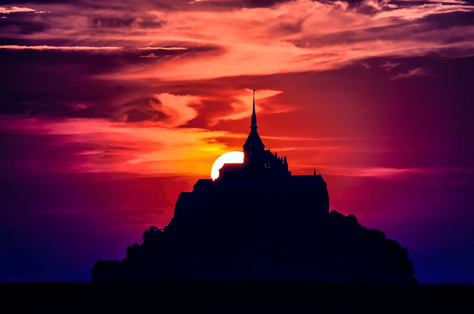 Silhouette of Mont Saint-Michel, France, at sunset. The dark shape of the island abbey is sharply contrasted against a vibrant, fiery sky with hues of red, orange, and purple. The setting sun is partially visible, creating a dramatic backlight and silhouette effect. Iconic French landmark, sunset landscape, Mont-Saint-Michel photography.