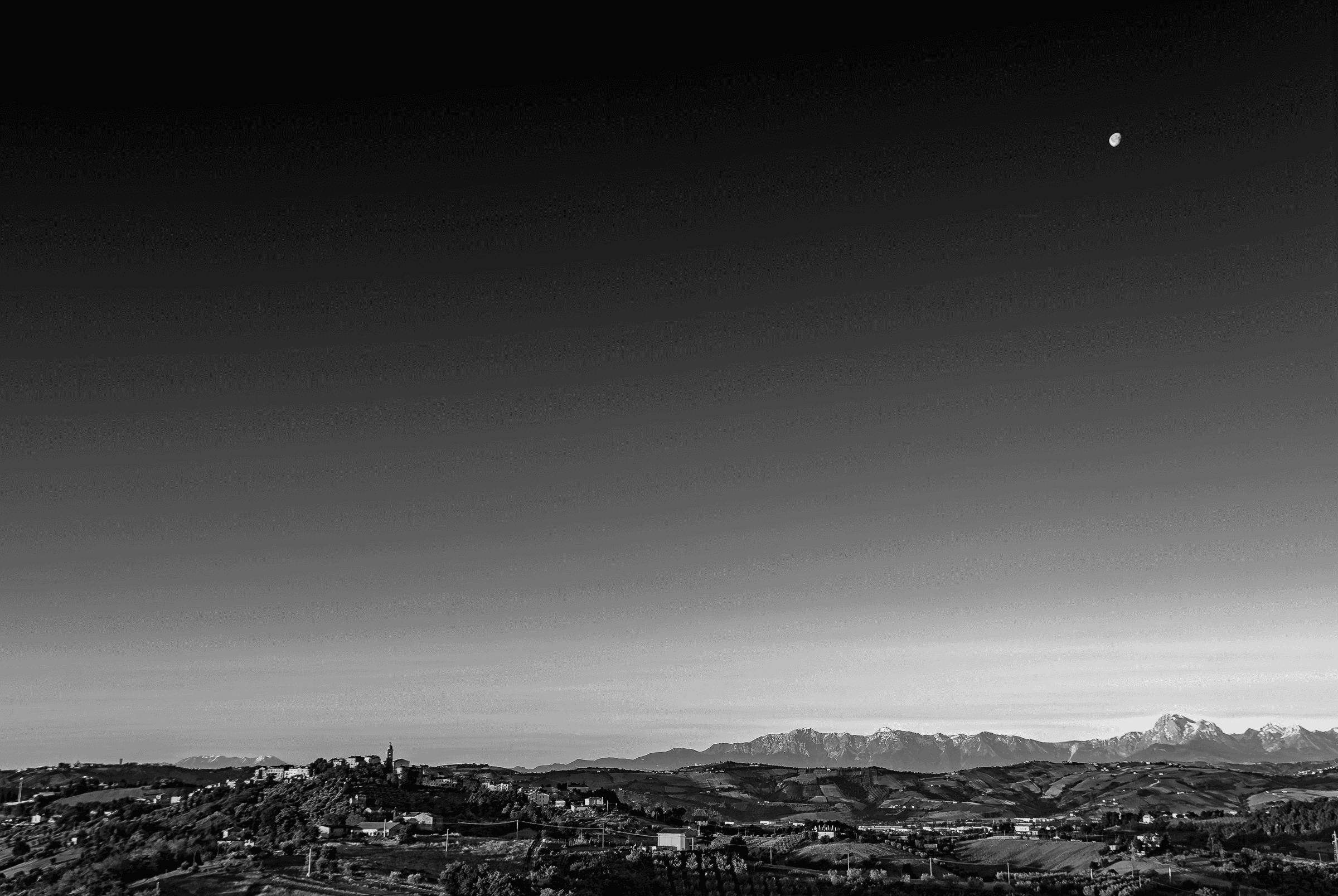 Black and white landscape photograph of a moonrise over the town of Monsampolo del Tronto, with the Gran Sasso mountain range in the background.