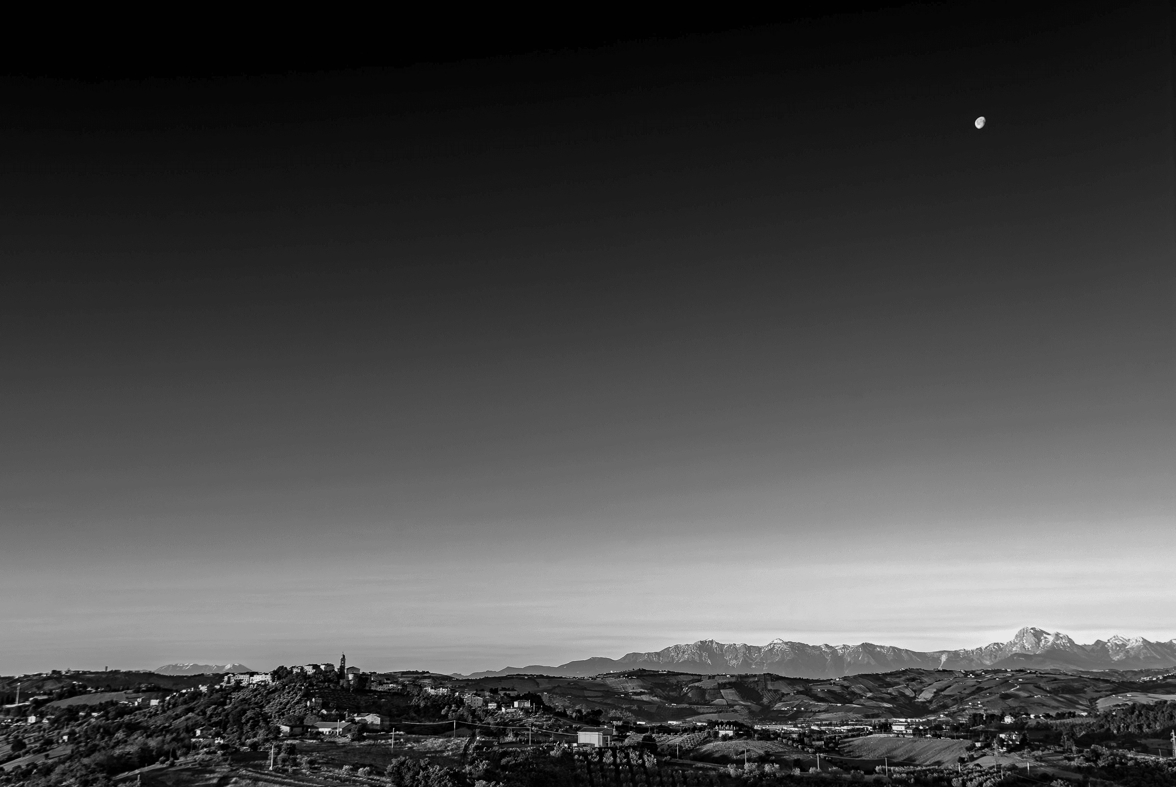 Black and white landscape photograph of a moonrise over the town of Monsampolo del Tronto, with the Gran Sasso mountain range in the background.